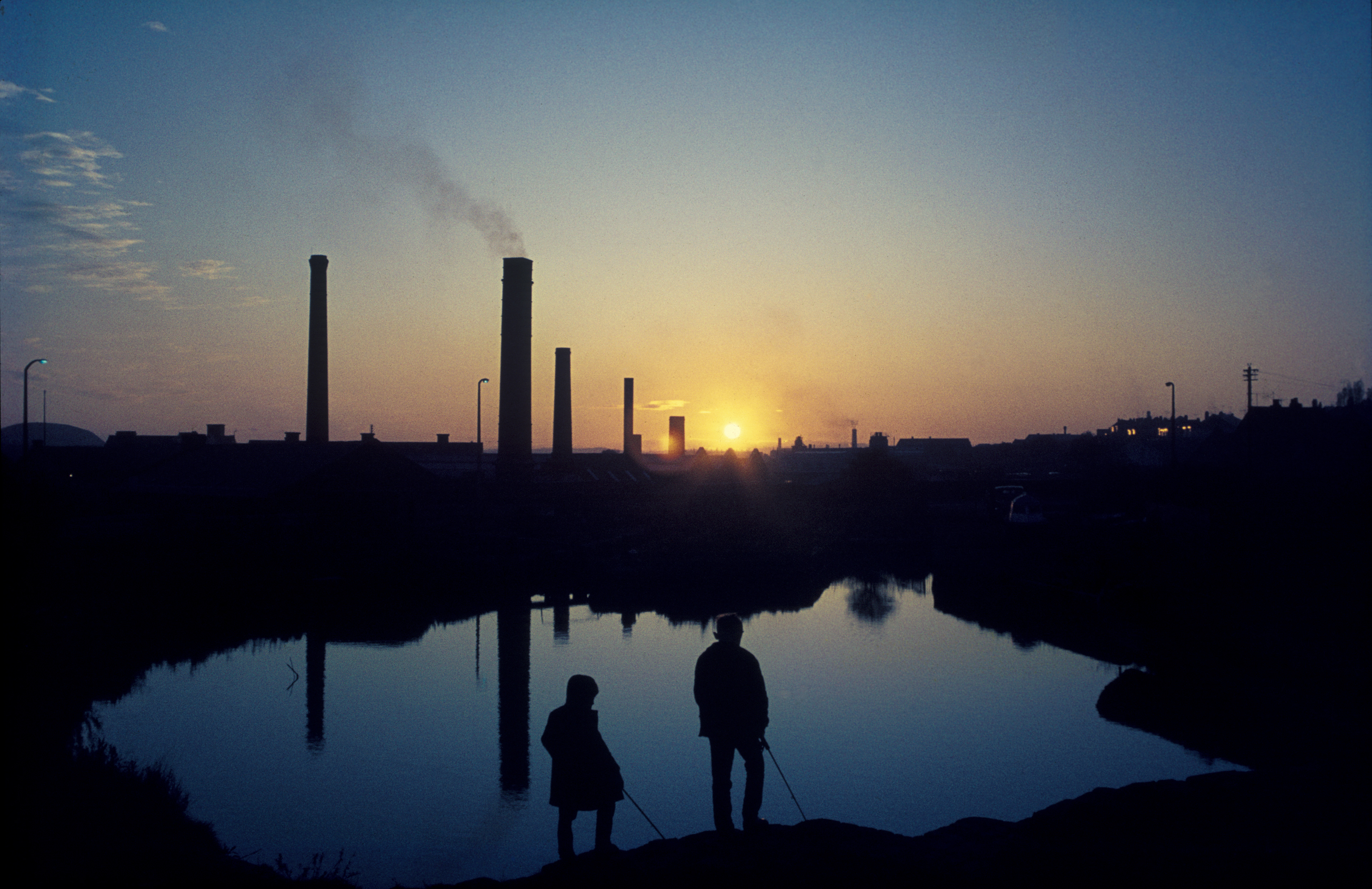 Silhouetted figures with fishing rods stand by a calm body of water at sunset. Smokestacks rise in the background, casting reflections on the water.