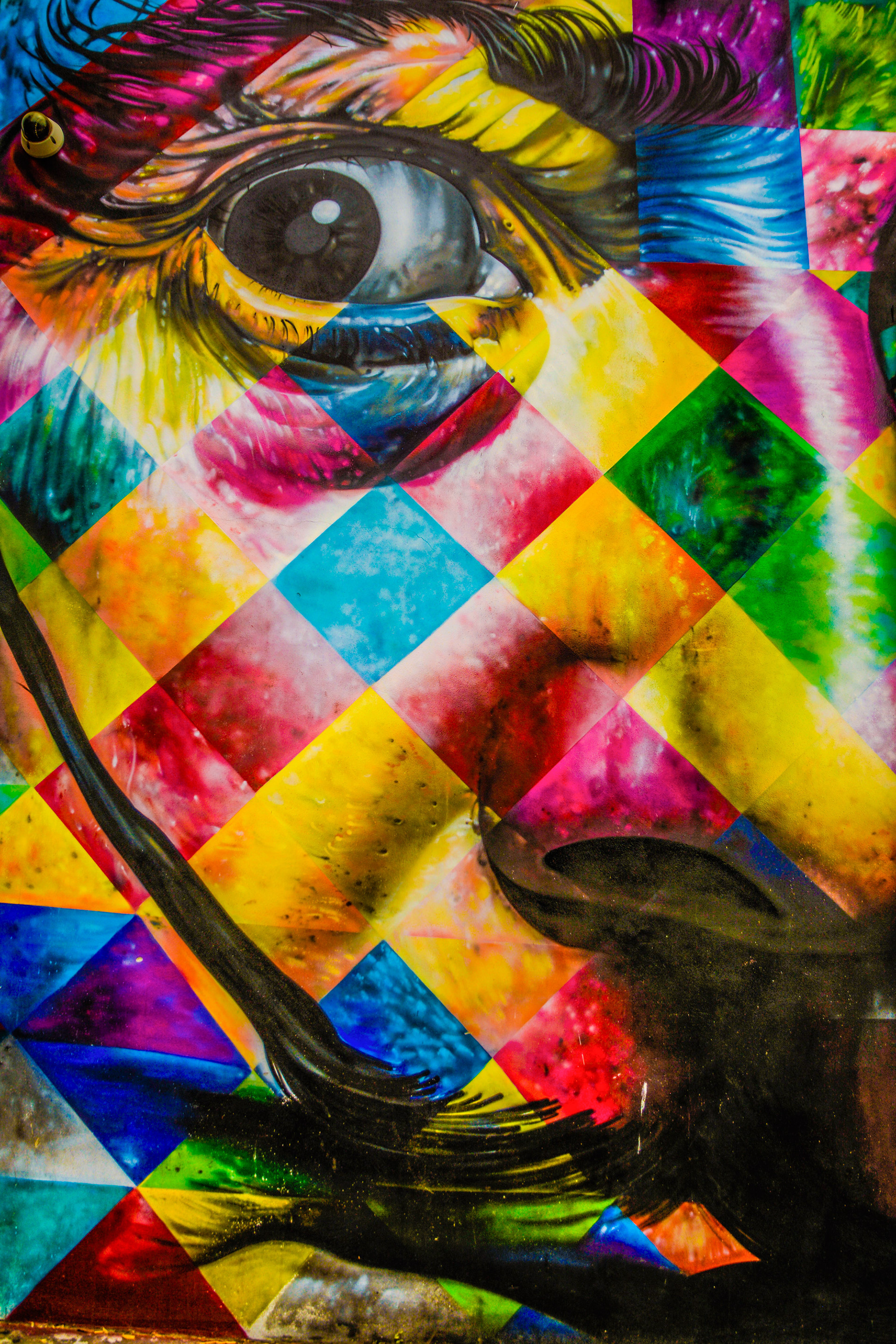 Colorful street art mural featuring a large, detailed Dali eye amidst a vibrant patchwork of geometric shapes and vivid colors, conveying a sense of intensity.
