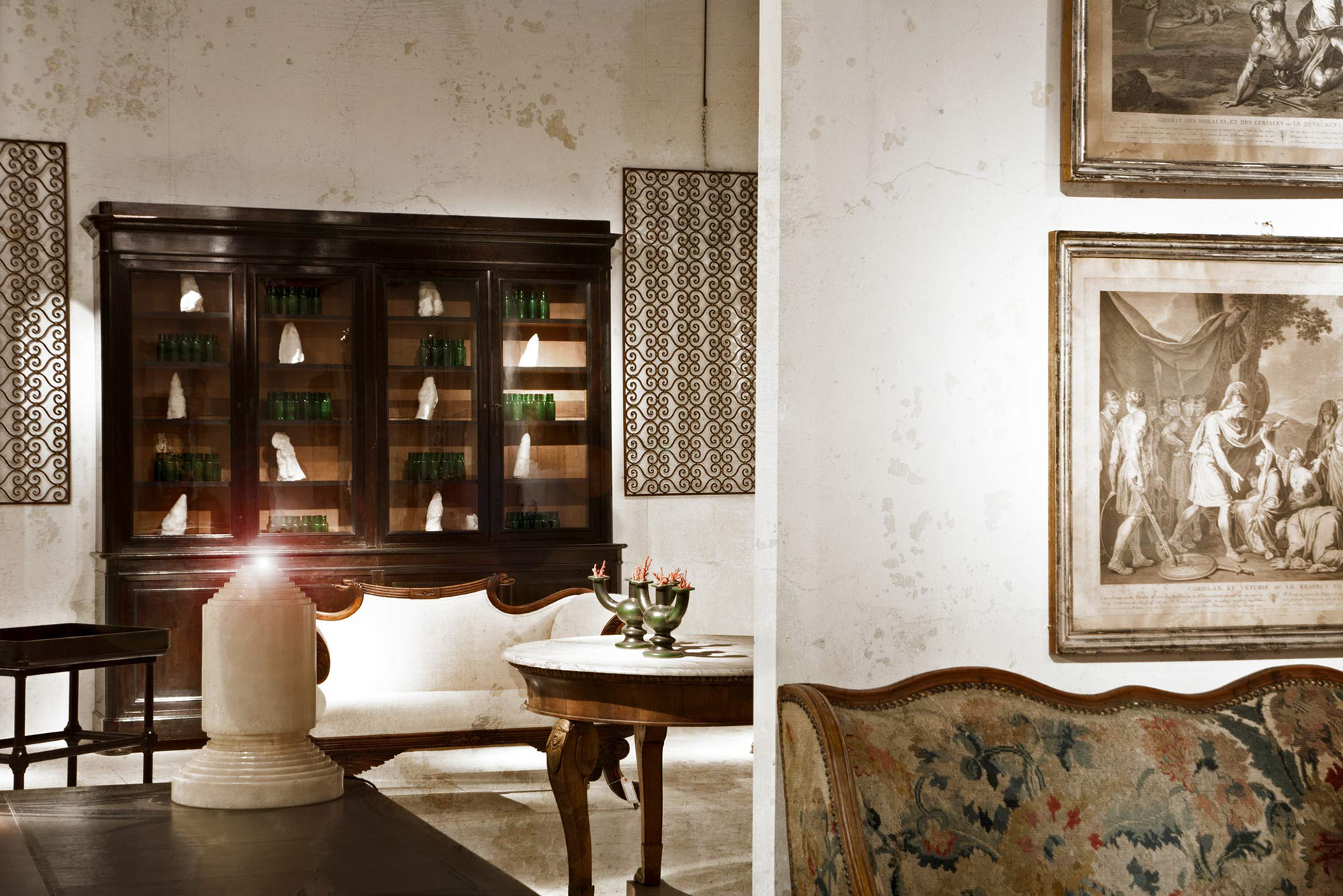 Vintage room with wooden cabinet displaying green bottles and white sculptures on shelves. Ornate wall art and patterned fabric sofa create an elegant, classic vibe.