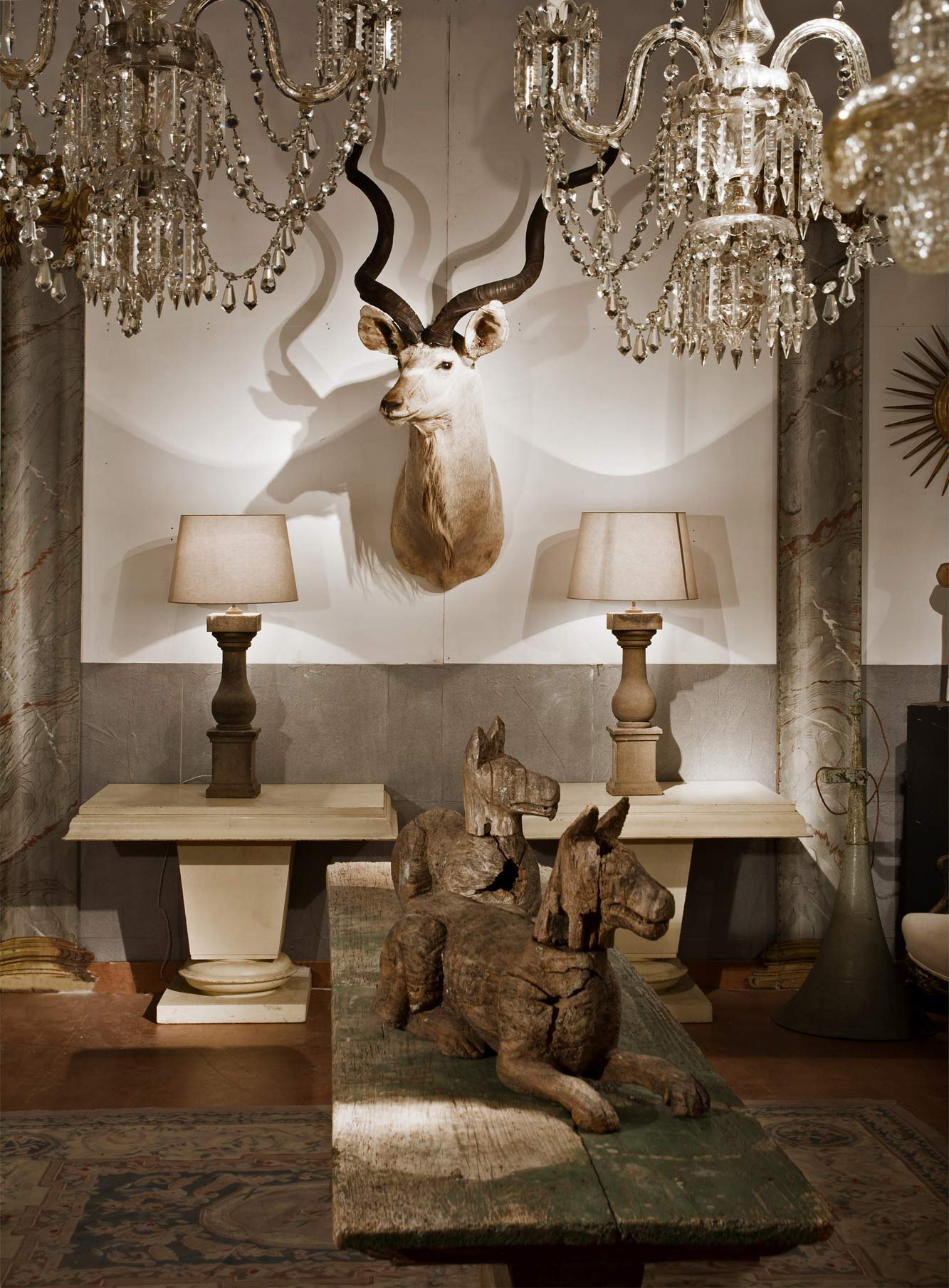 Rustic room with mounted antelope head, two table lamps, wooden horse sculptures, column, sunburst mirror, and classical statue, exuding an antique vibe. photographed in the cotswolds by simon donnelly