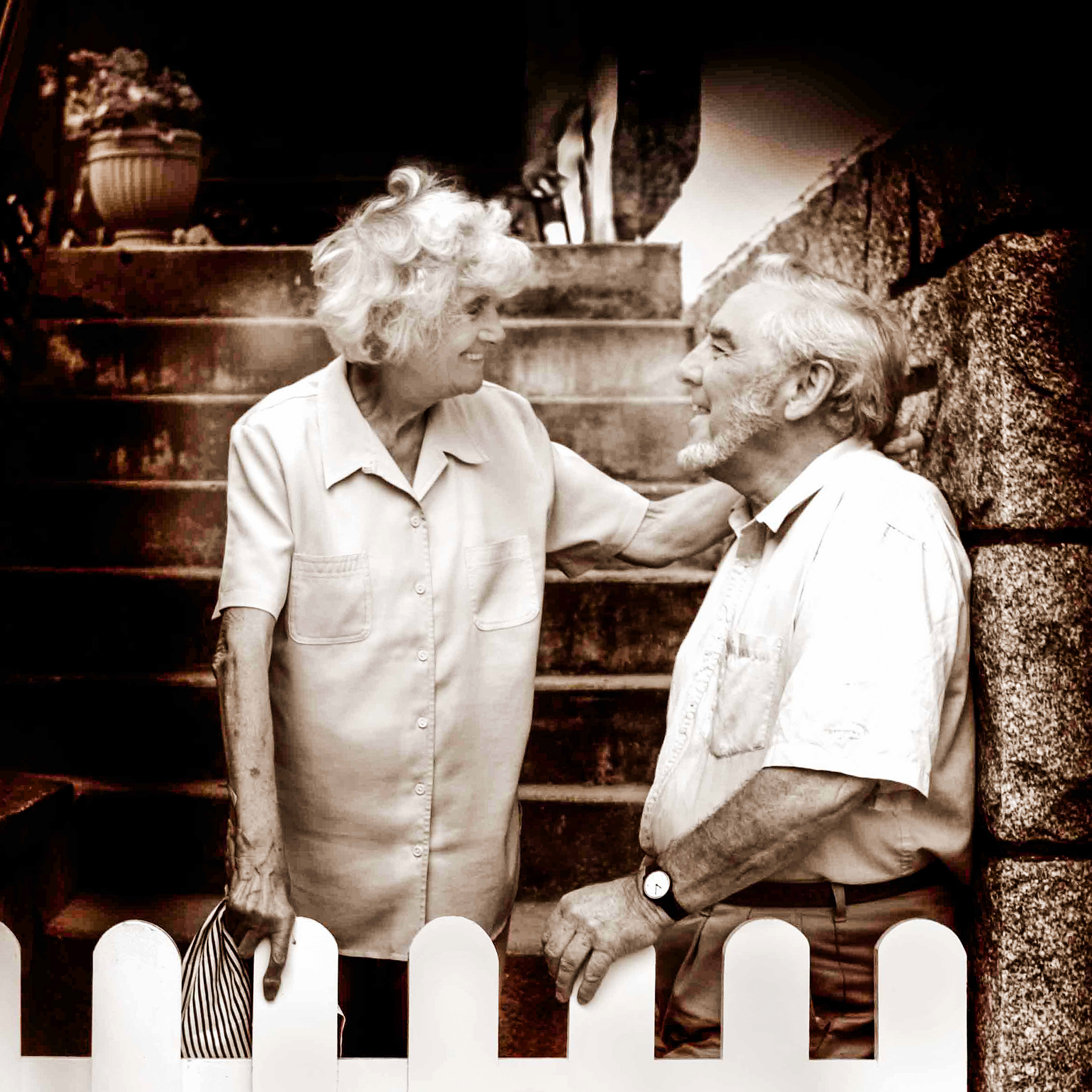 Elderly couple smiling warmly at each other near stone steps and a potted plant; woman gently holds man's shoulder, conveying affection and nostalgia.