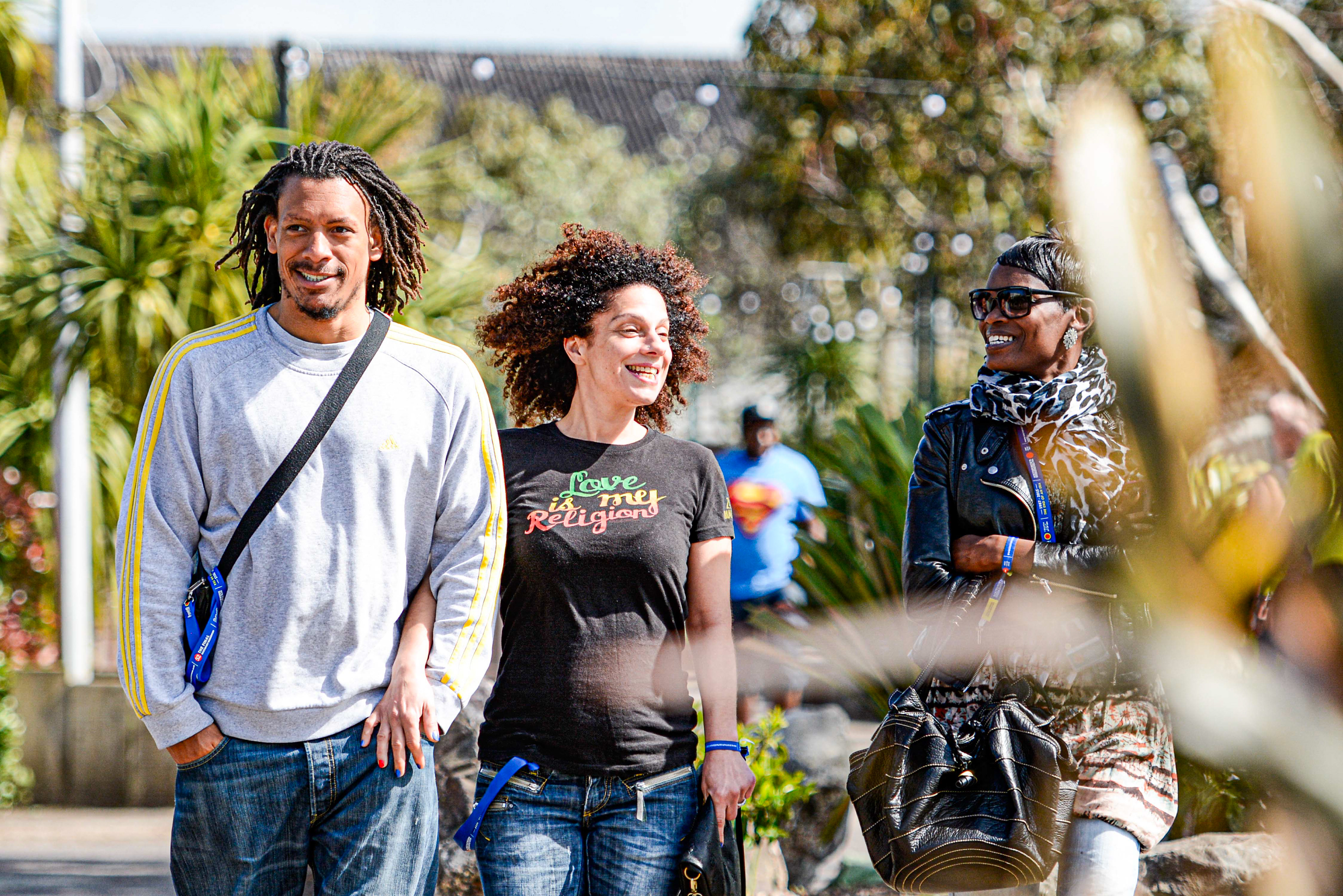 Three people walk outdoors smiling and chatting. The person on the left wears a gray sweater, the middle wears a shirt saying "Love is my Religion," and the one on the right wears sunglasses and a scarf. Bright, sunny day with greenery in the background.