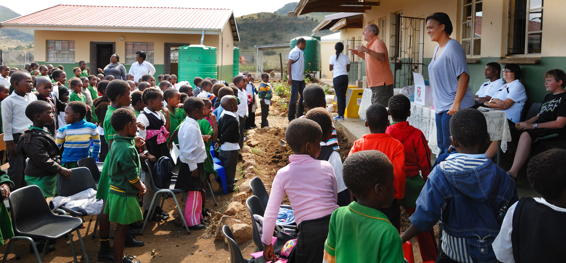 Children in green and colorful uniforms stand outside a school building with adults interacting. The scene is lively and engaging, conveying a joyful atmosphere.