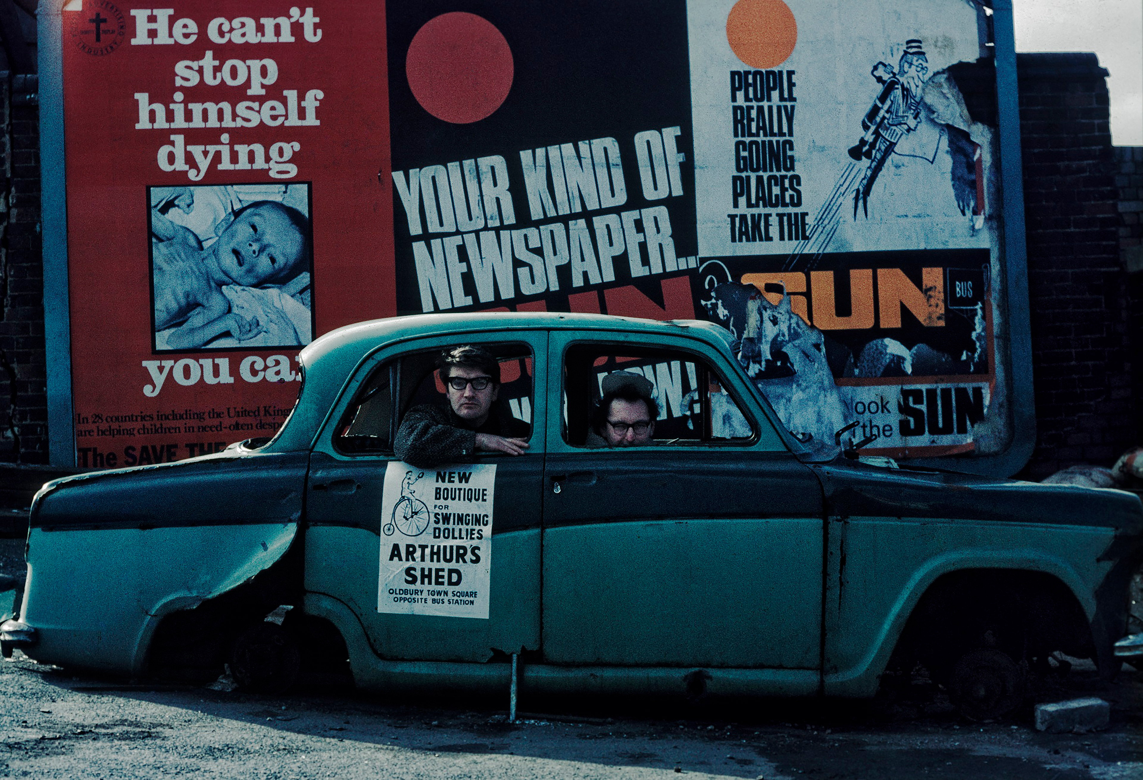 Two men sit in a rusty, abandoned car in front of vibrant, peeling billboards. The scene conveys decay and urban neglect, with a sombre atmosphere.