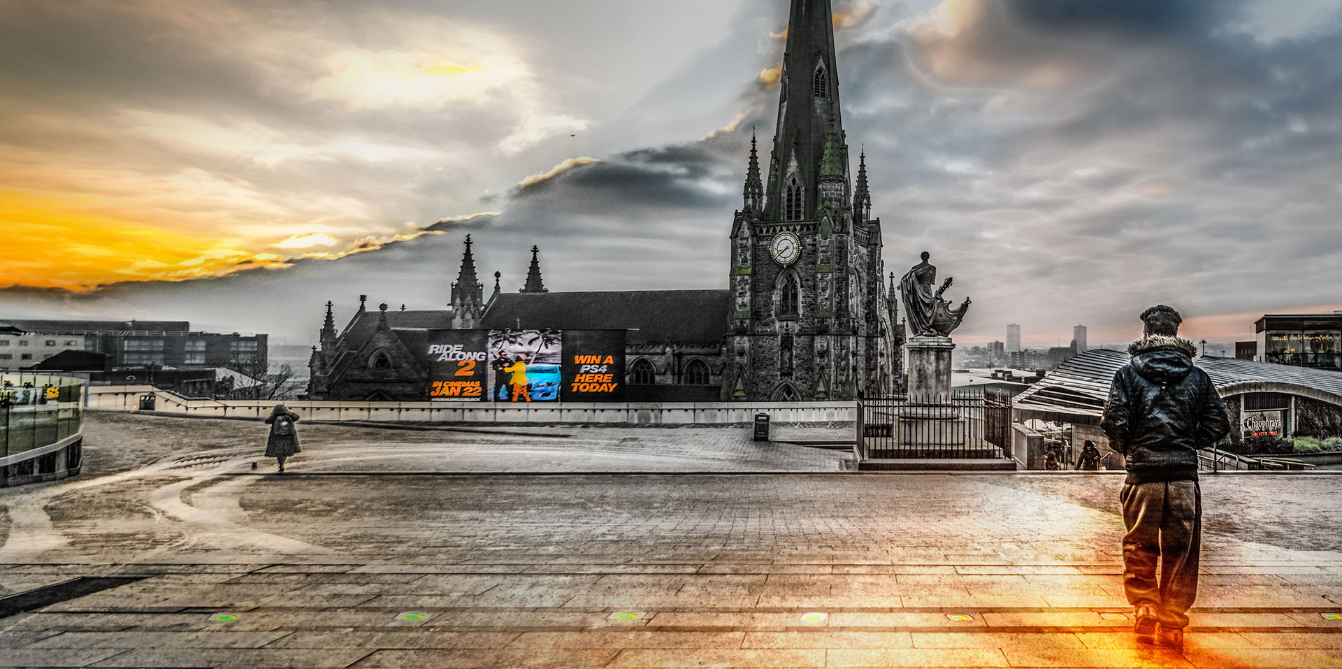 A person in a dark jacket stands on a wet street, facing a historic church with a clock and spires. Bright digital billboards are vivid against a dramatic, cloudy sunrise. Professional advertising photographer Birmingham