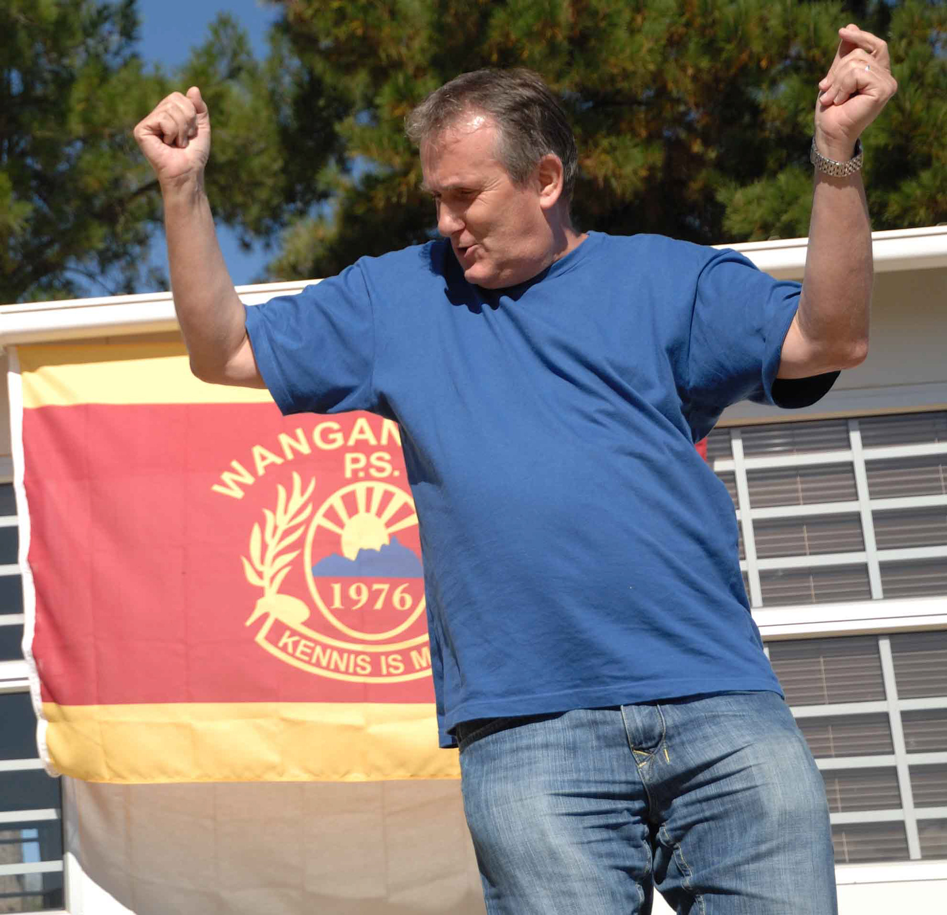 A man in a blue shirt raises his fists in a celebratory manner, standing in front of a red and yellow banner with an emblem, under a clear blue sky.