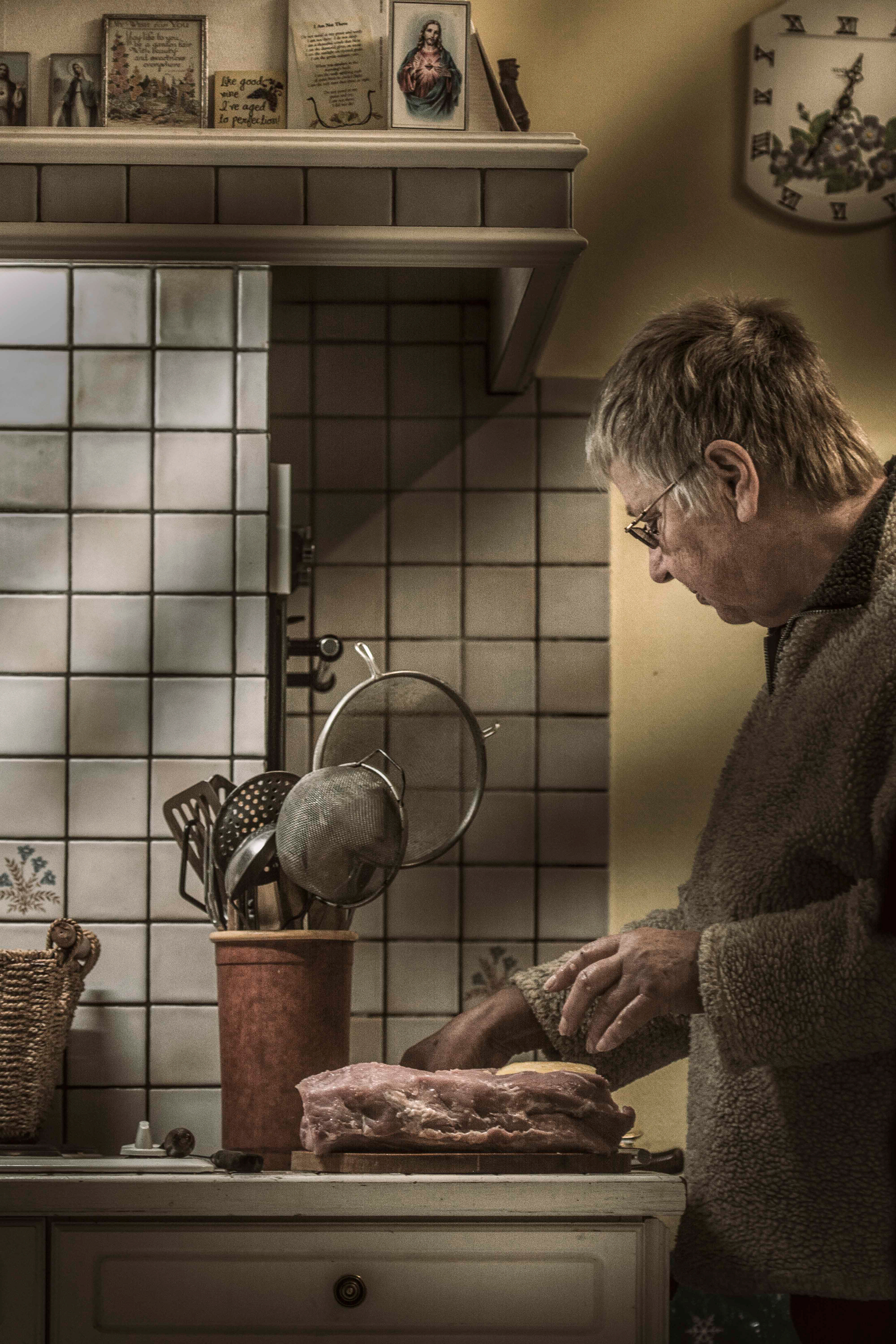 An elderly person in a cozy sweater prepares food in a kitchen with tiled walls. Utensils are stored in a terracotta holder nearby, creating a warm, homey atmosphere.