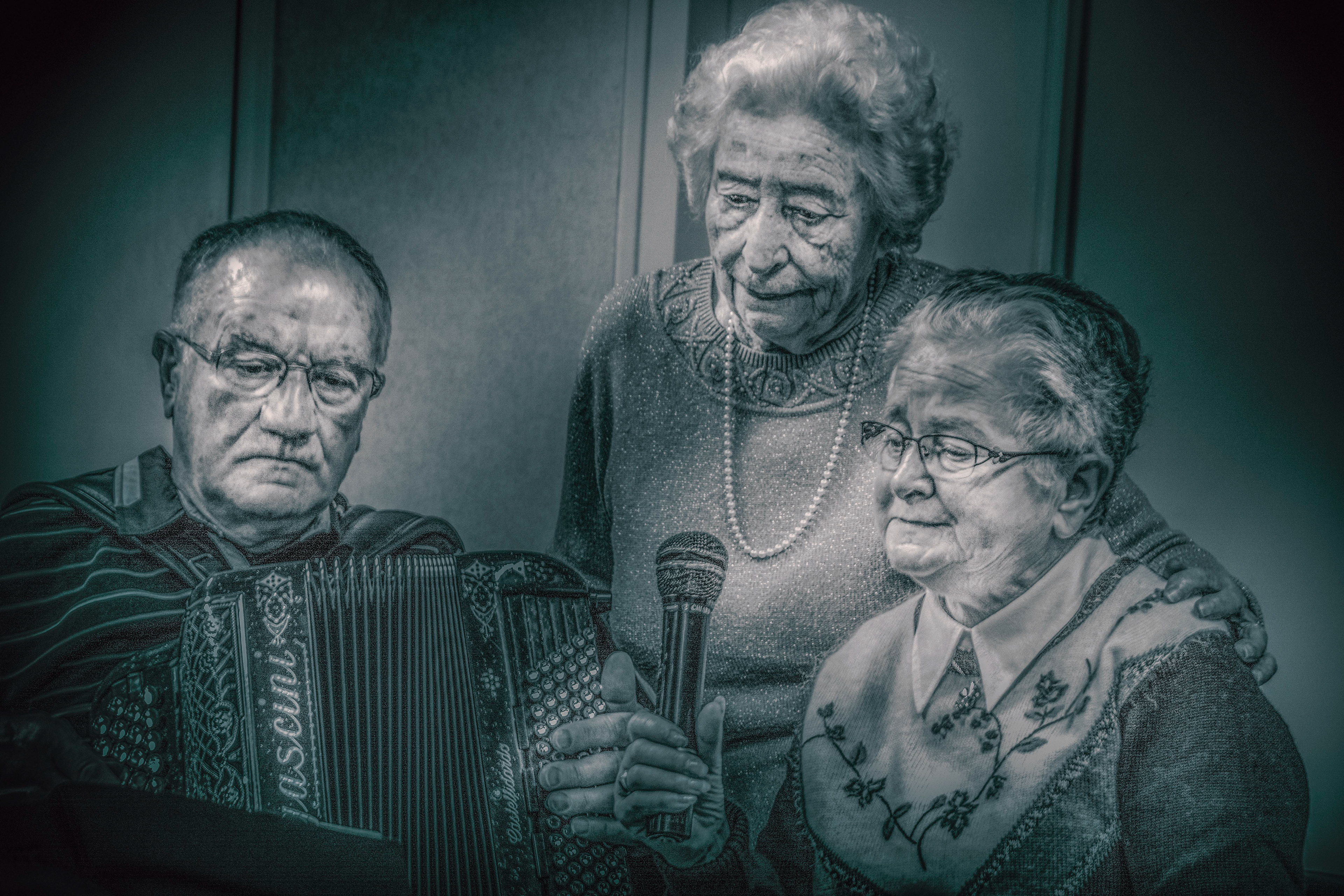 Three elderly people gather, one holding an accordion, another with a microphone, sharing an intimate, nostalgic moment. Monochrome tone.
