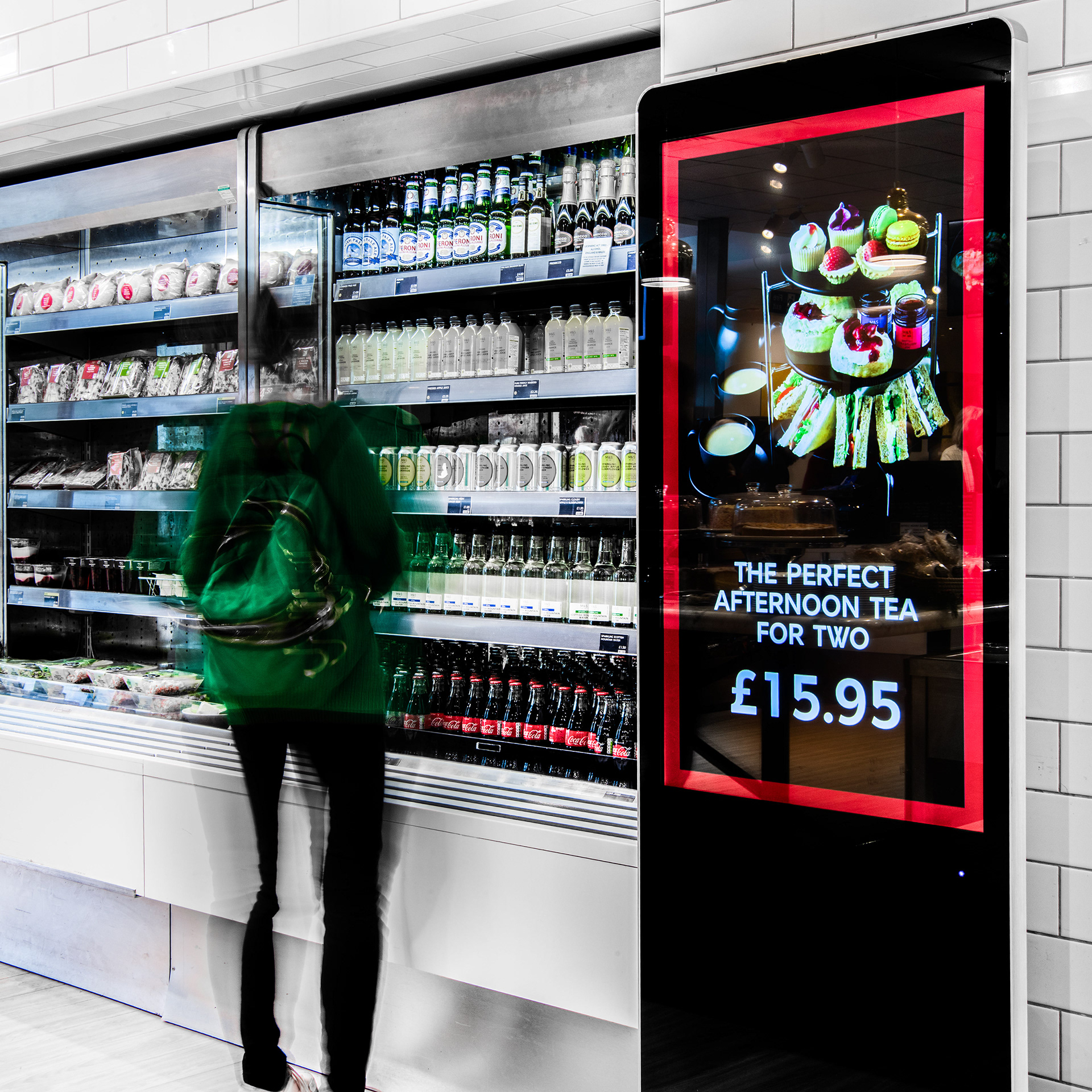 A person wearing a green jacket and backpack stands in front of a grocery fridge with drinks and sandwiches. A bright ad promotes afternoon tea for two.