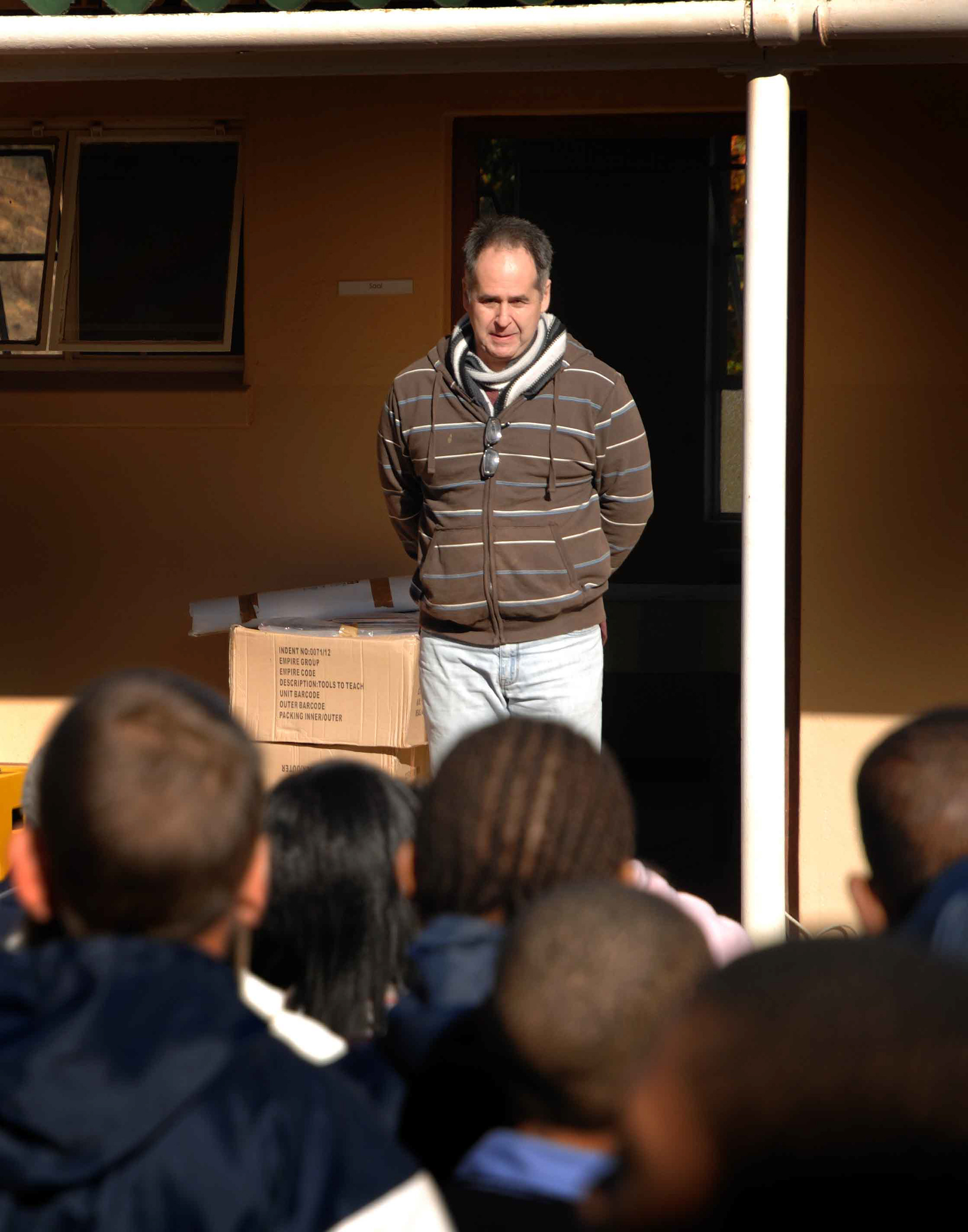 A man in a striped hoodie and scarf stands outside a building, facing an audience. He appears contemplative. A cardboard box is on his left.