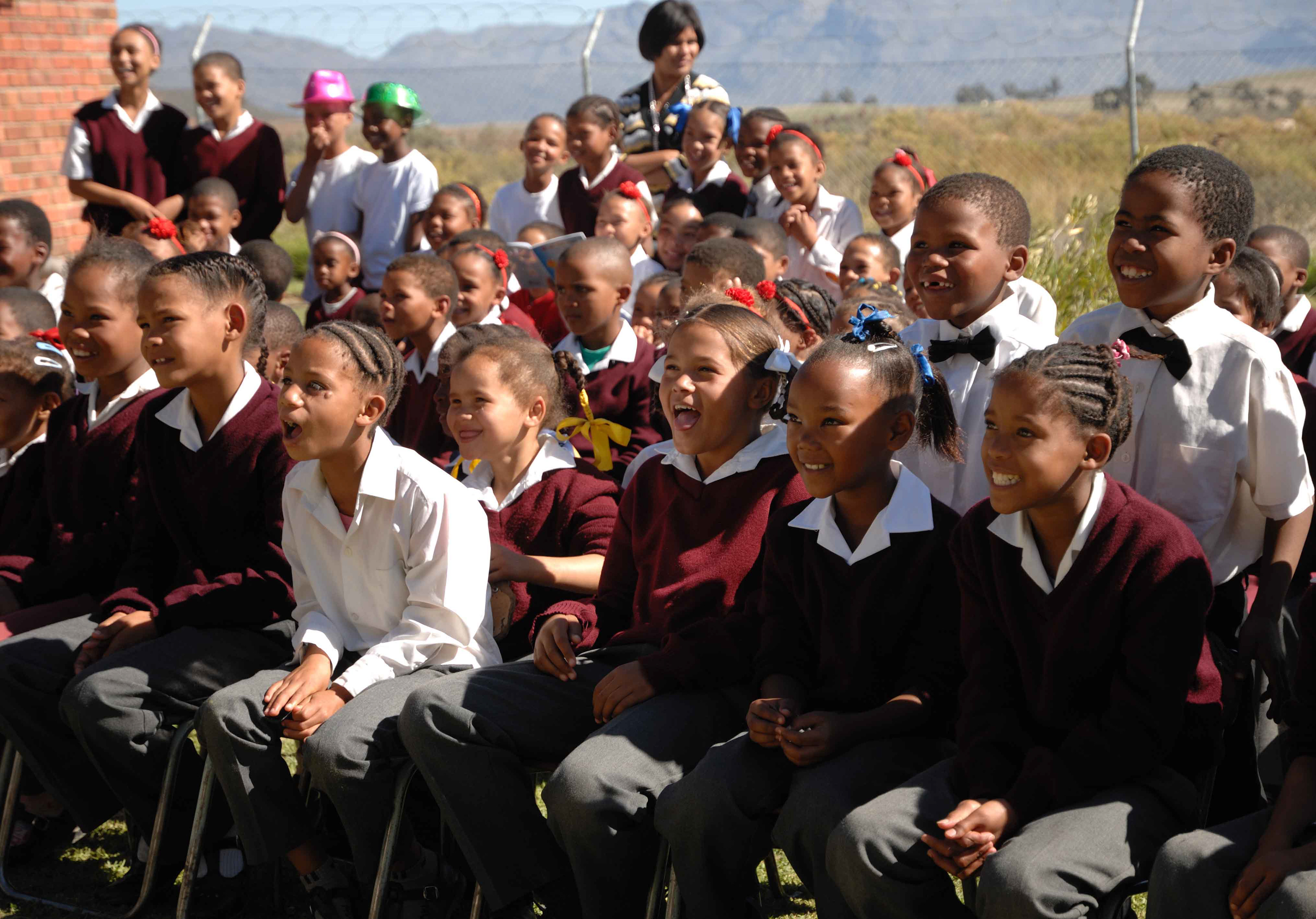 A joyful group of schoolchildren in uniforms, sitting outdoors, smiling and engaged. Bright sunlight enhances the lively and cheerful atmosphere.