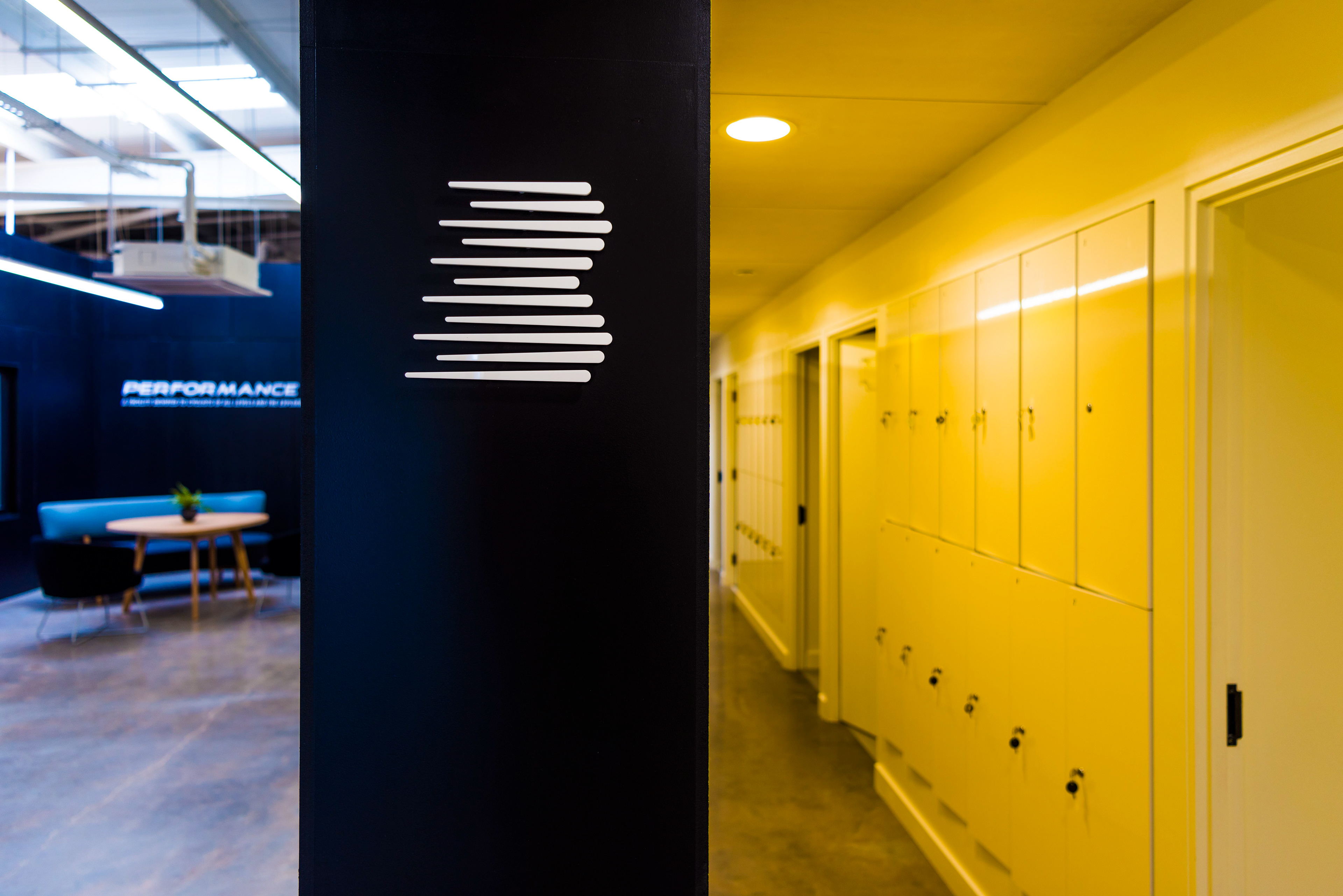 A modern office hallway with a black wall featuring white, horizontal lines. To the right, yellow lockers line the corridor. The atmosphere is clean and sleek.