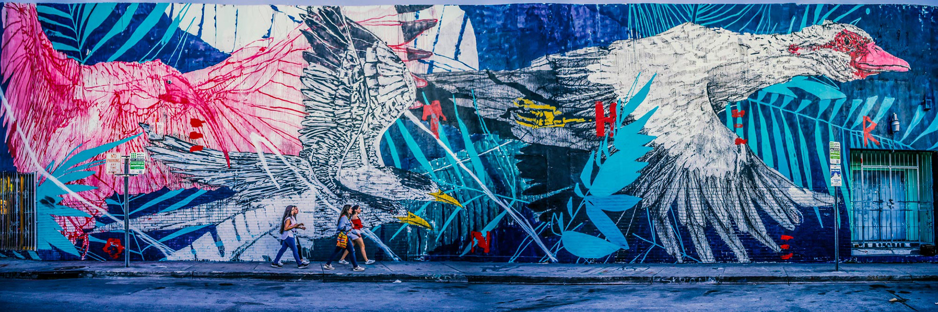 Street art mural of vibrant pink and white birds with bold lines against a blue leafy background. Three people walk by, adding movement. Urban, colorful, dynamic.