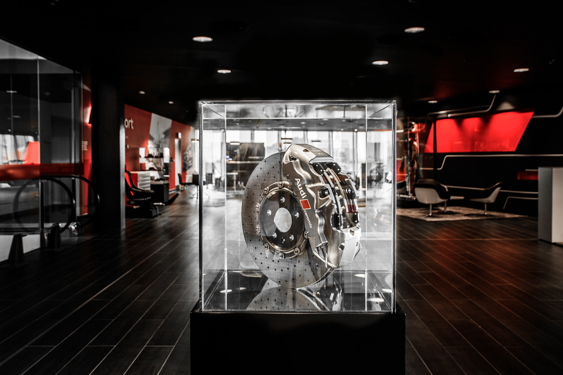 Exhibit of an Audi ceramic brake disc and caliper encased in glass. The setting is a modern showroom with dark, sleek decor, evoking sophistication. Lighting by Simon Donnelly Photographer