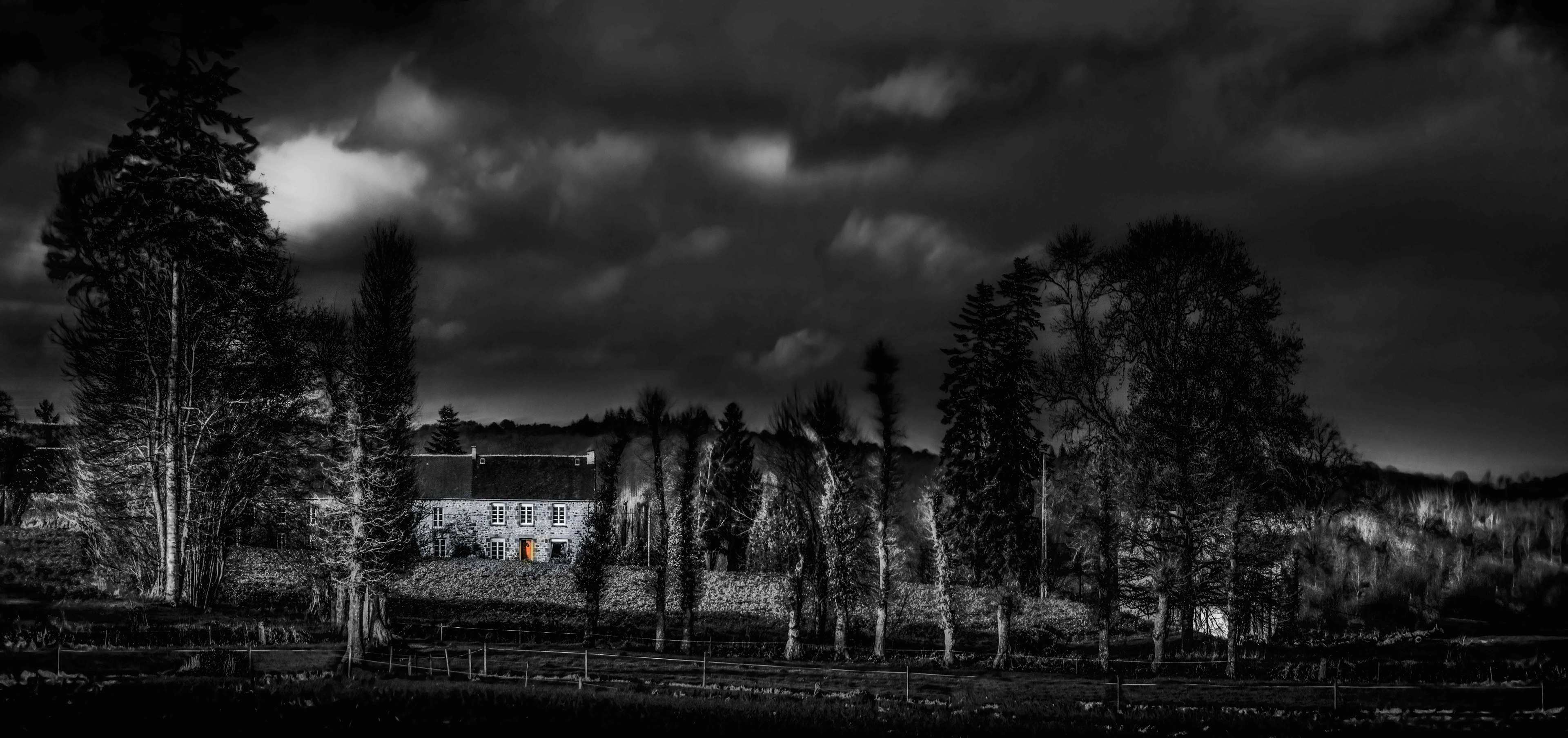 A dimly lit stone house with a glowing orange door amidst bare trees under a moody, cloudy night sky, creating an eerie yet inviting atmosphere.