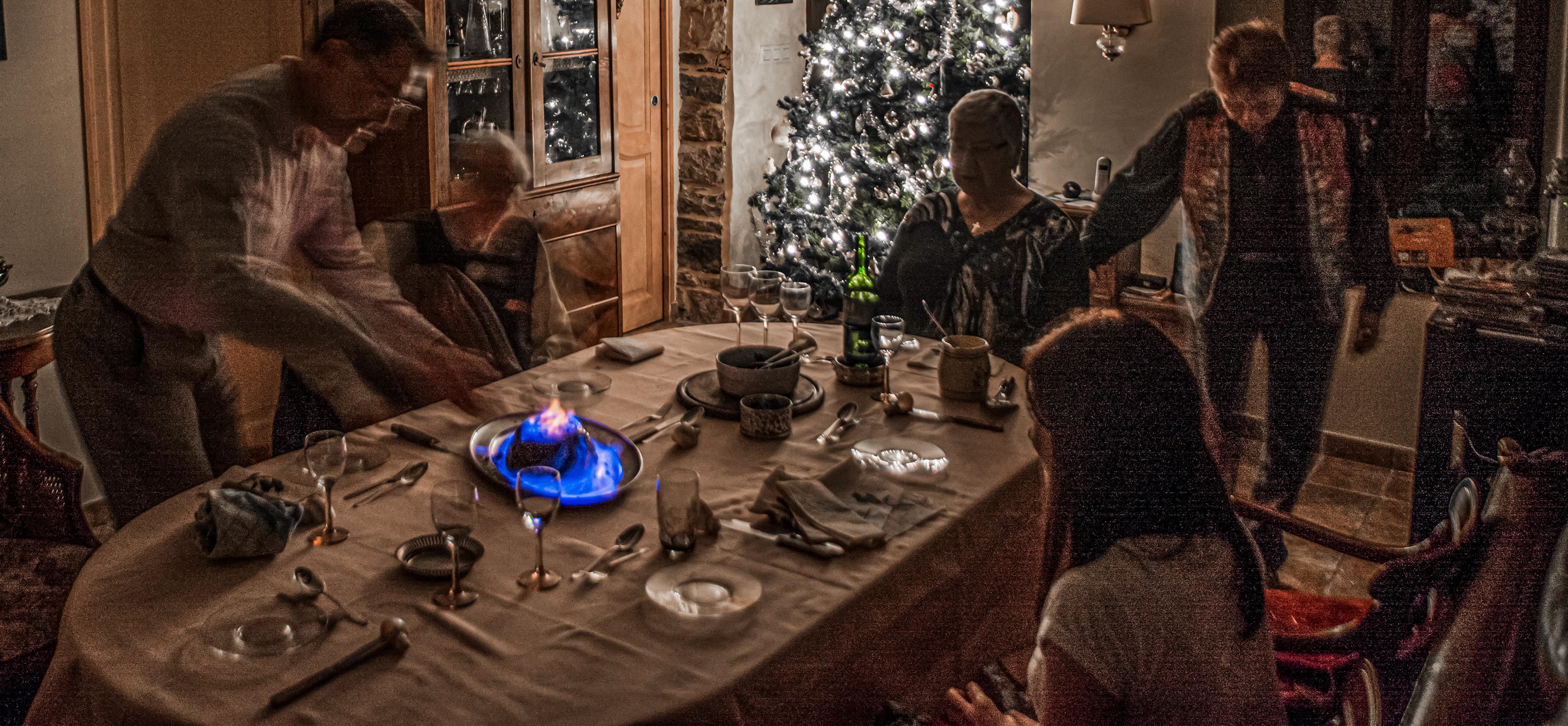 A festive dining scene with six people around a table, featuring a blue-flamed dessert. A decorated Christmas tree glows warmly in the background.