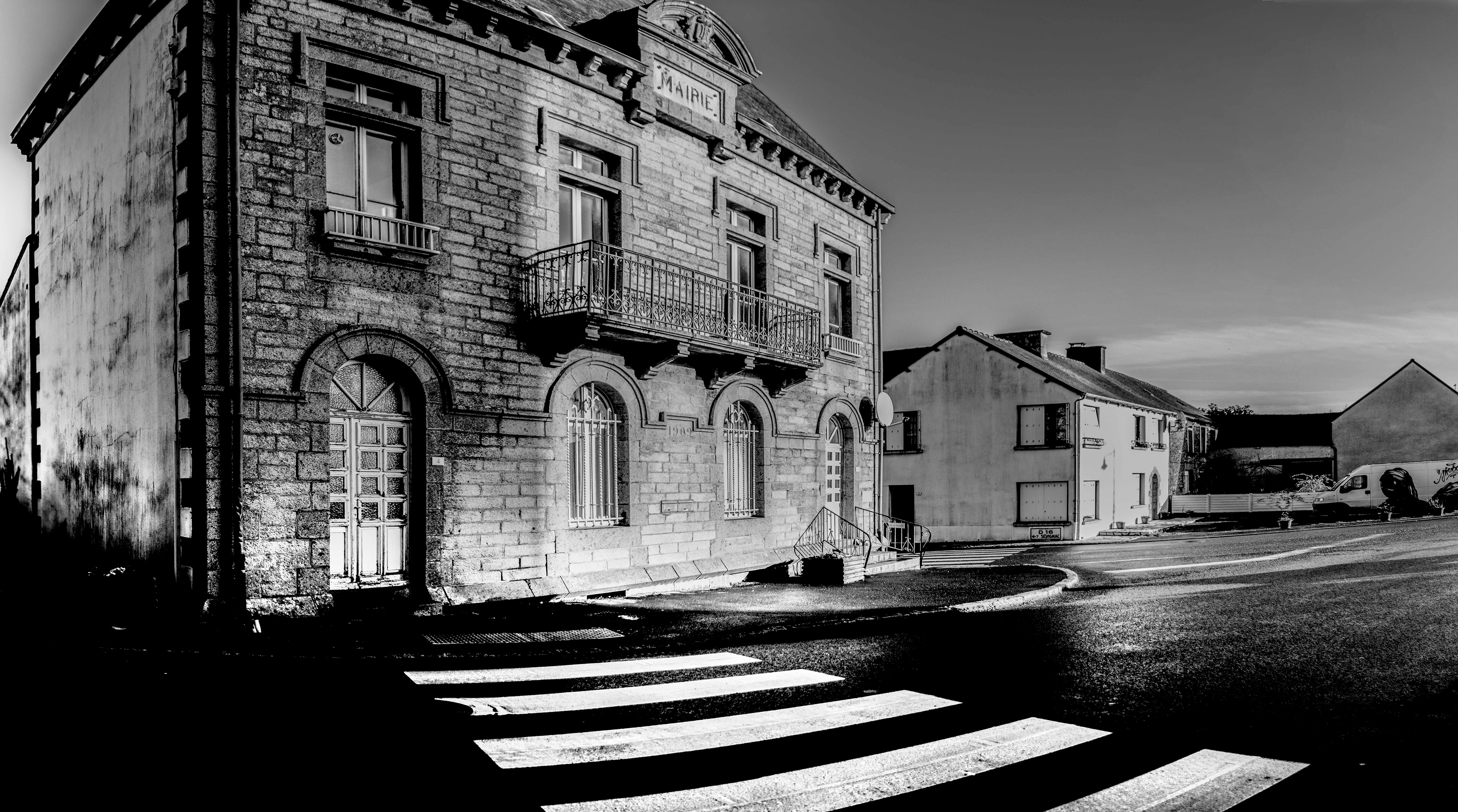 Black and white photo of a historic stone building marked "Mairie" with arched windows and balcony, alongside quiet street and crosswalk shadows.