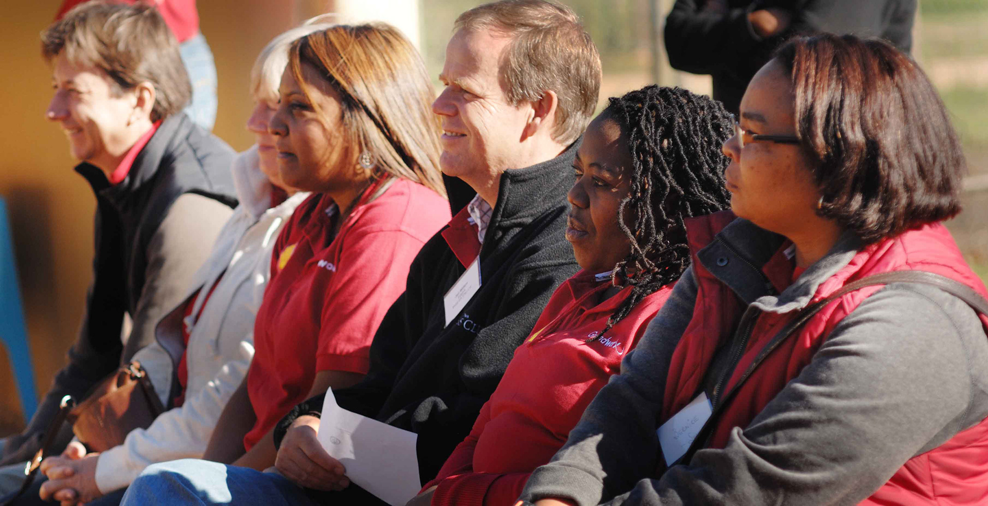 A diverse group of people sit in a row, wearing casual jackets, smiling and focused, suggesting warmth and camaraderie in an outdoor setting
