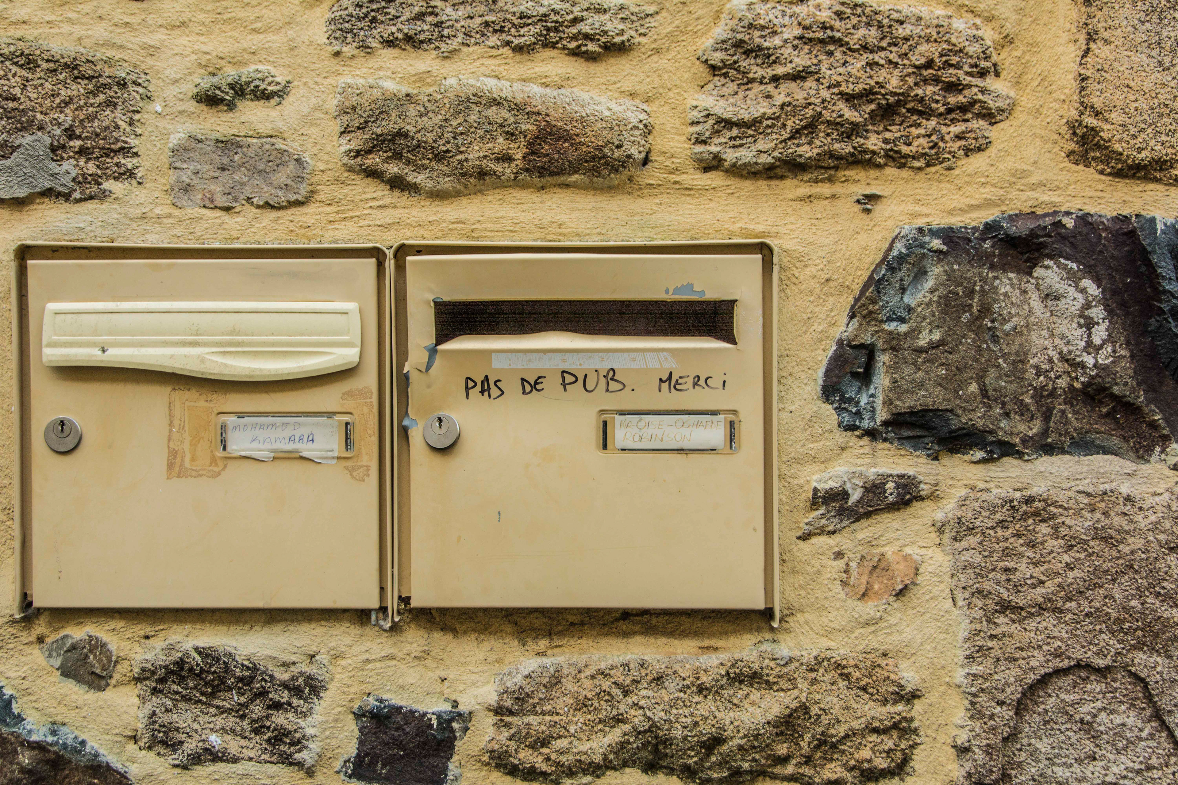 Two beige mailboxes are mounted on a rustic stone wall. One has a note reading "Pas de pub. Merci," conveying a polite request for no ads.