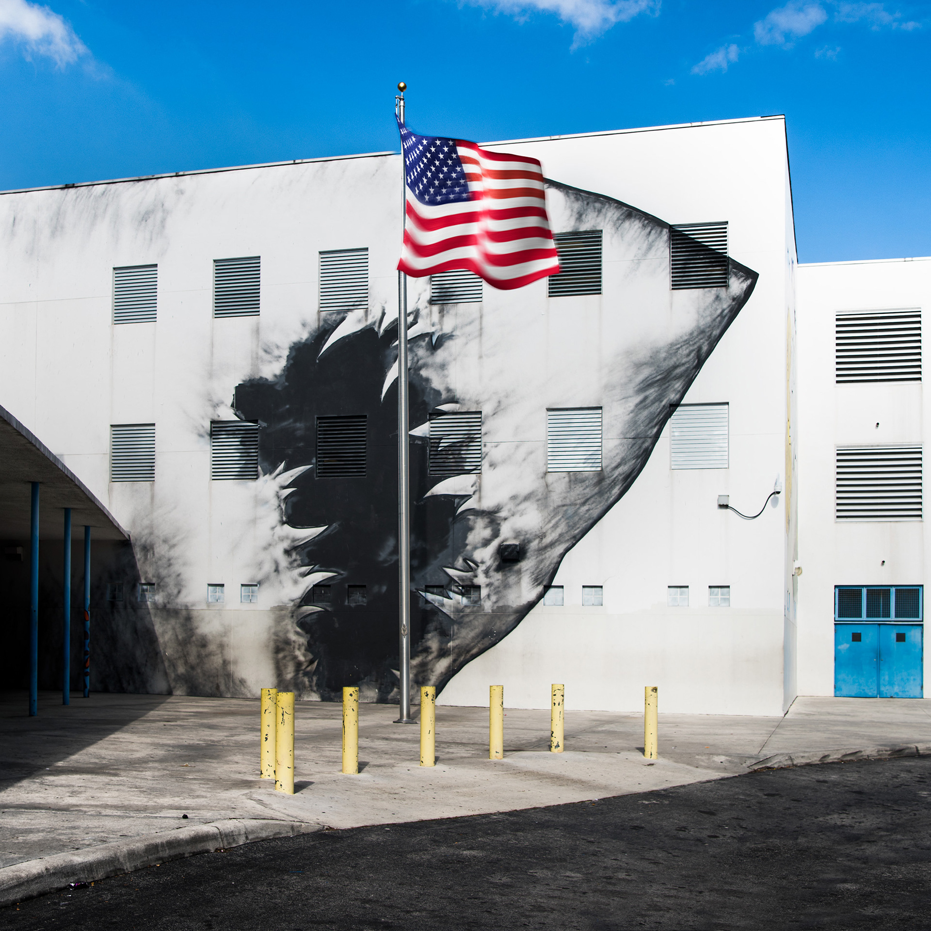 A U.S. flag waves in front of a modern white building with a dramatic black mural resembling a shark. The scene is set under a bright blue sky.