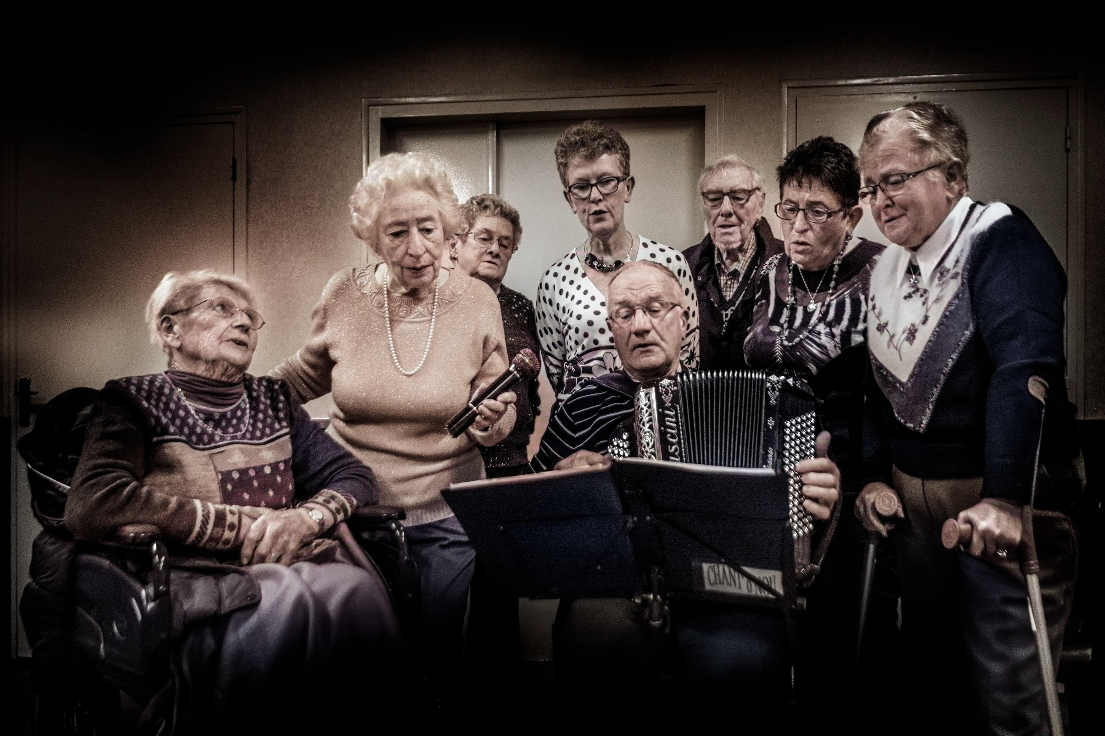 A group of elderly people singing together; one plays the accordion, another holds a microphone. They appear focused and joyful in a cozy room.