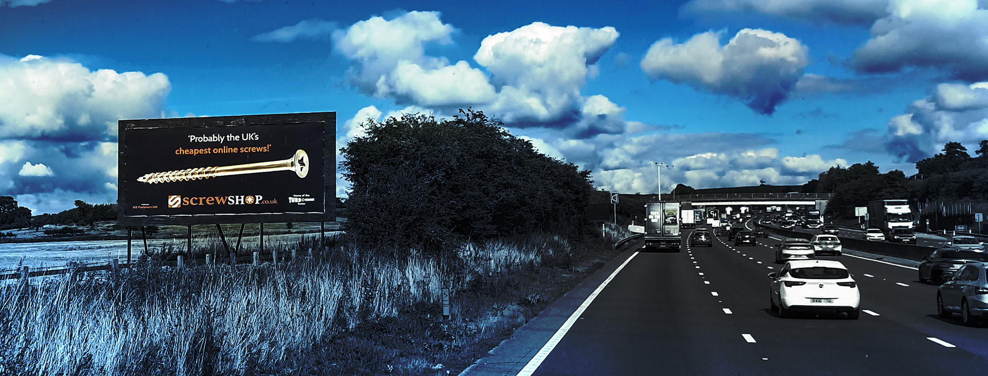 Highway scene with traffic under a blue, cloud-filled sky. A large billboard on the left advertises screws, setting an industrial and commercial tone.