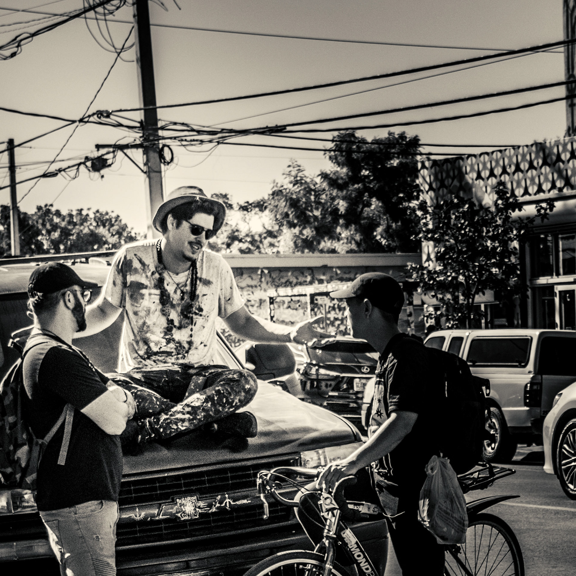 A man in a hat and sunglasses sits on a car hood, animatedly talking to two friends, one on a bike, in a lively urban setting. Energetic atmosphere. Simon Donnelly, Birmingham Photographer