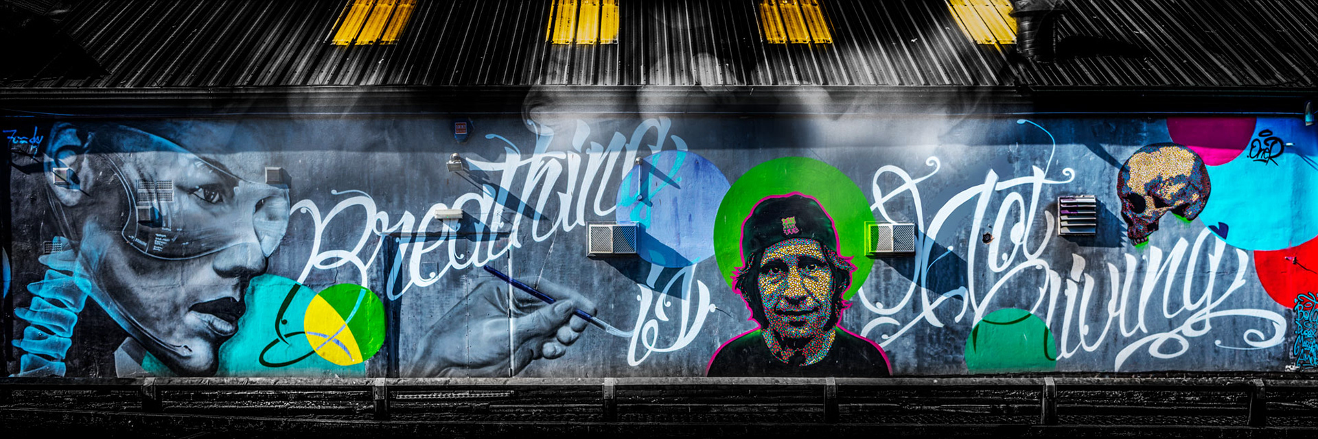 Simon Donnelly photographing graffiti at Liverpool Disco Festival,. Rodney Mullen USA skate boarding legend. Street art mural with a large face in profile wearing goggles on the left, colorful abstract patterns, text "Breathing, Not Giving," and a stylized skull on the right. Atmosphere is bold and urban.