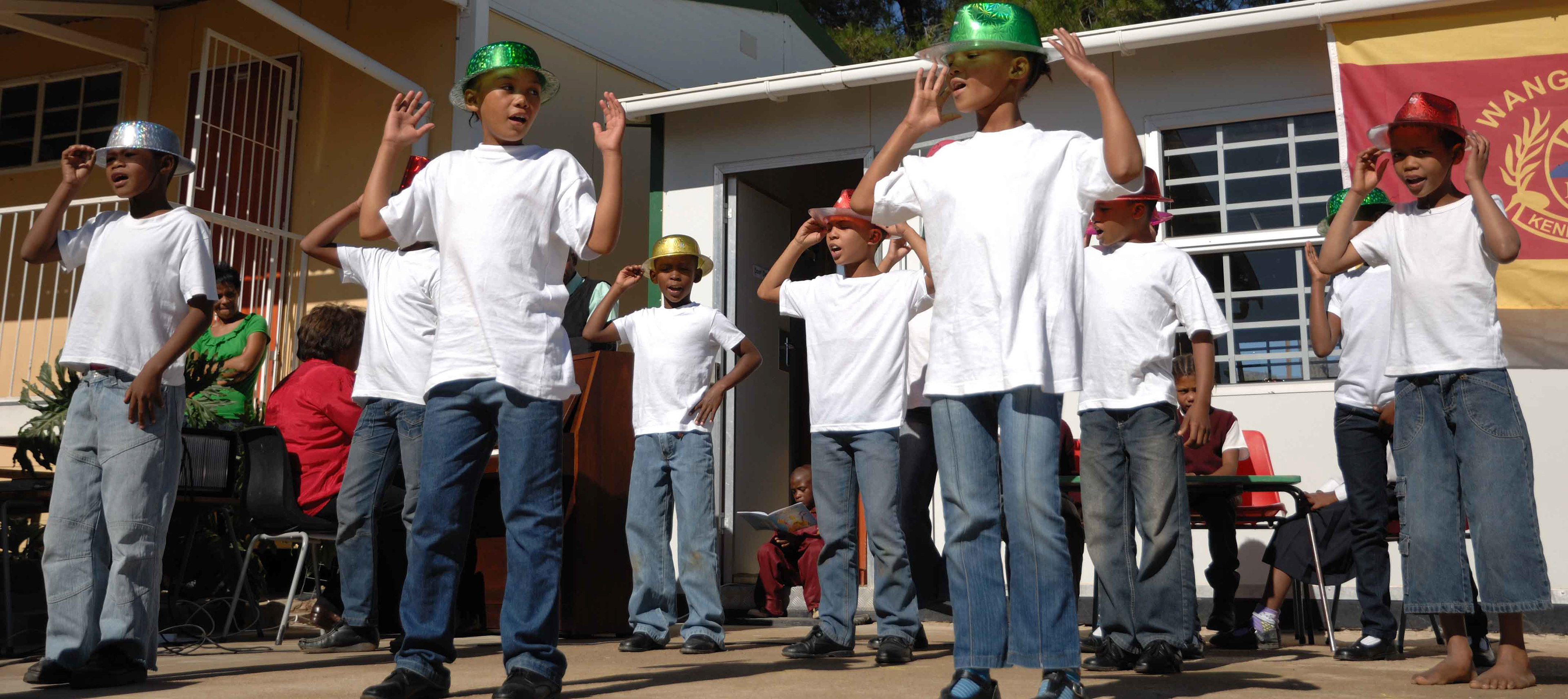 A group of children in white shirts and jeans perform outside. They wear colorful shiny hats and strike animated poses. The setting is lively and festive.