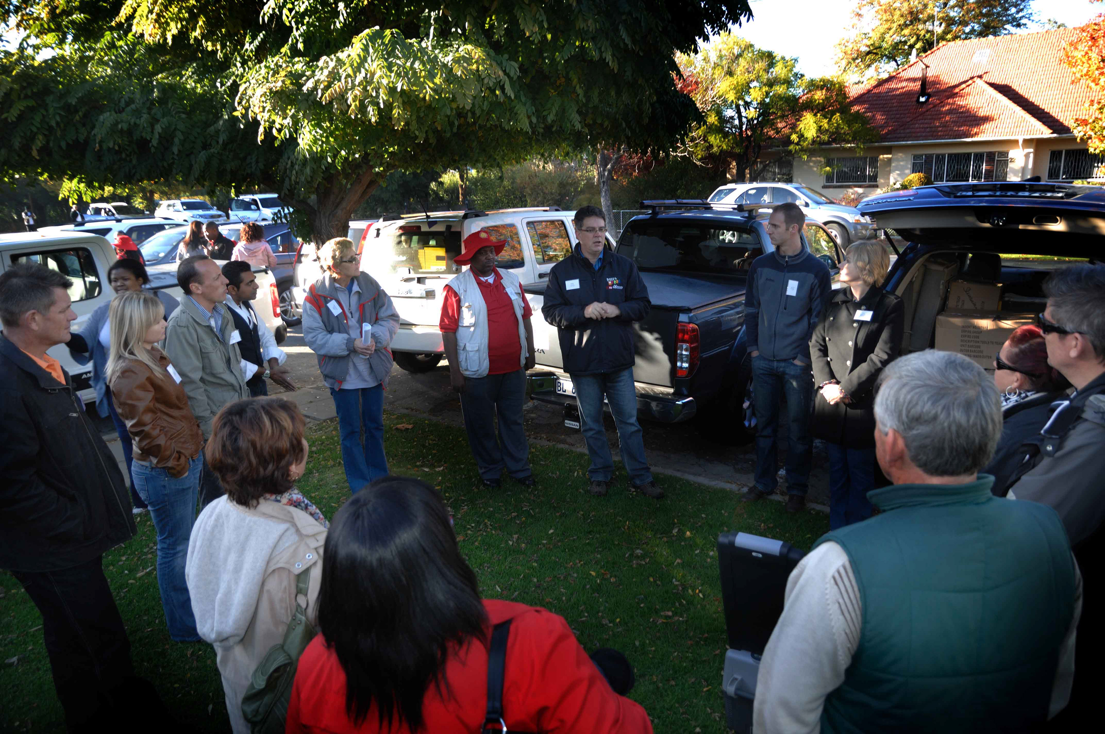 A group of people gather outdoors in a circle on a grassy area, attentively listening to a person speaking. Cars and trees are visible in the background.
