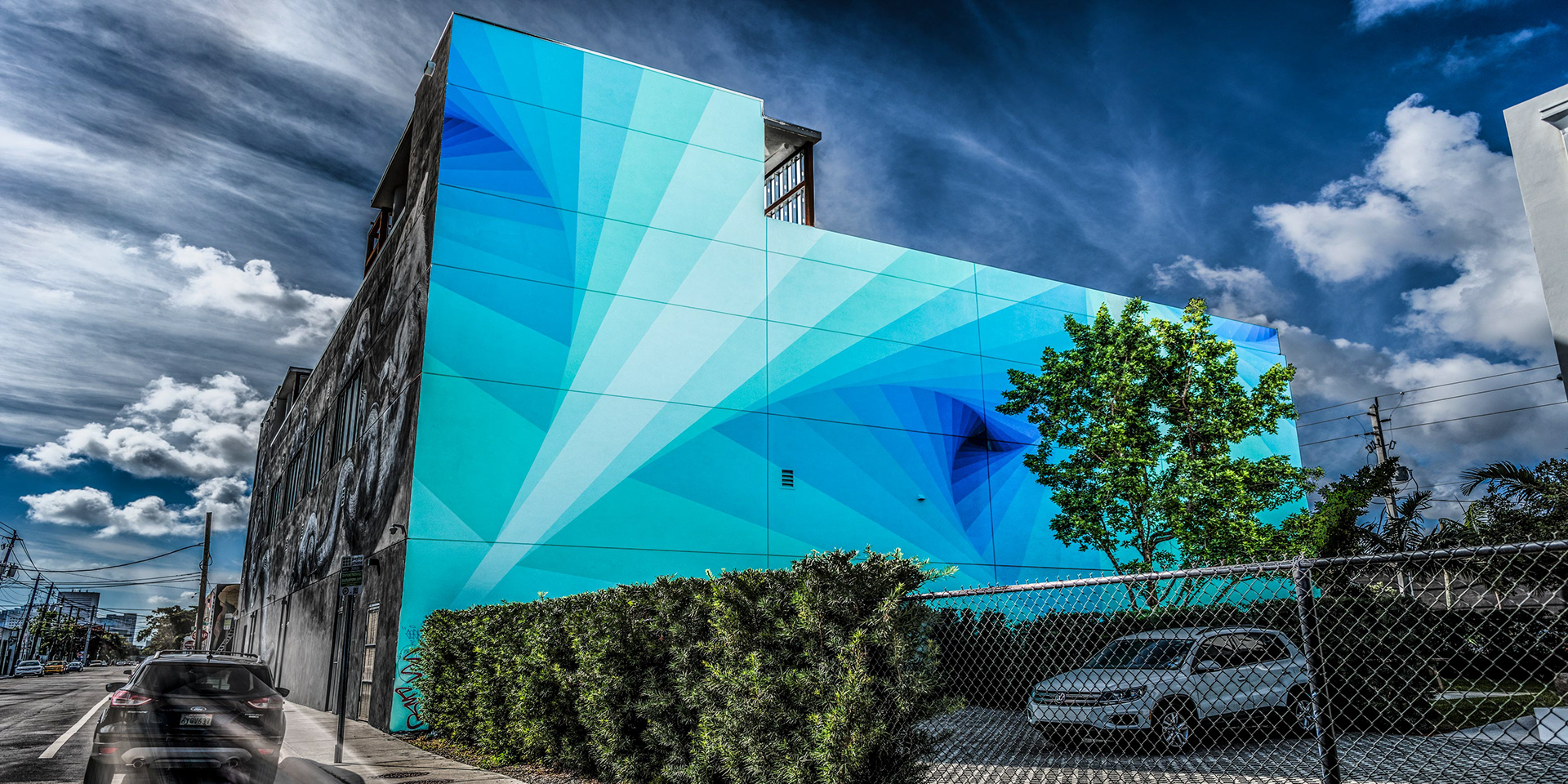 Street scene with a vibrant geometric mural in shades of blue on a building, next to a road with cars. The sky is dramatic with clouds.