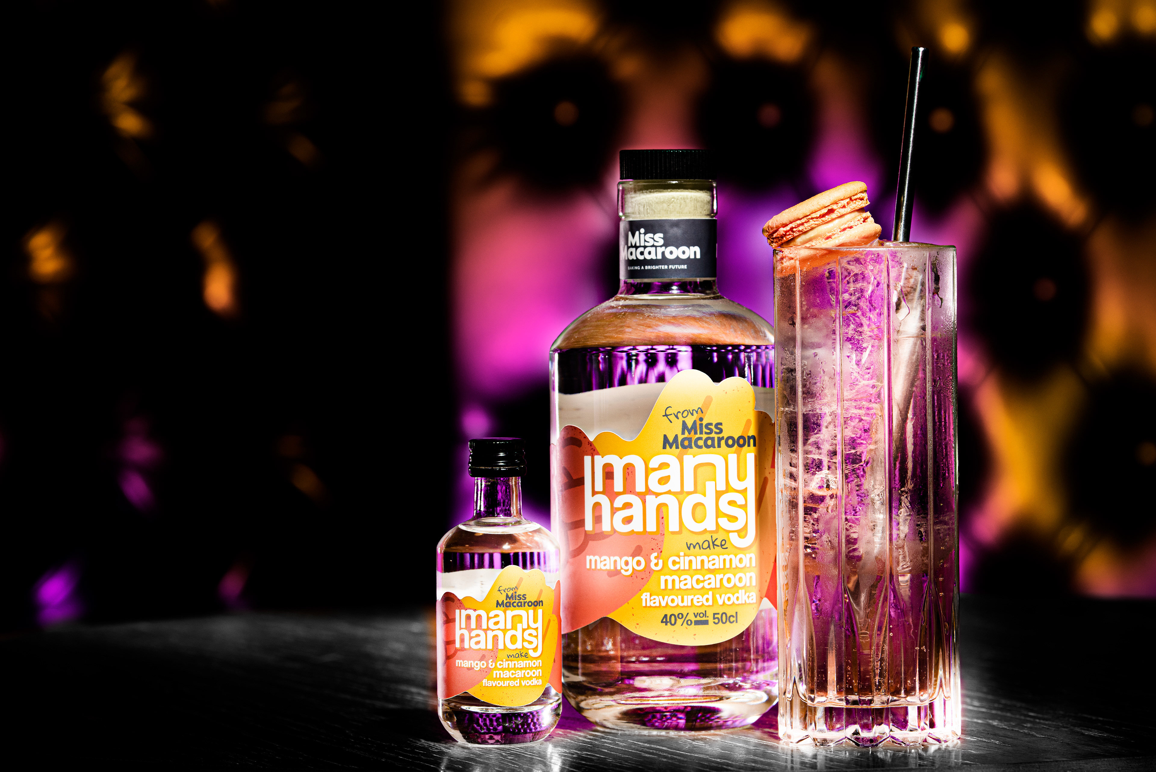 Bottles of Miss Macaroon's mango and cinnamon macaroon vodka stand on a table with a tall cocktail glass. Vibrant purple and orange bokeh background.