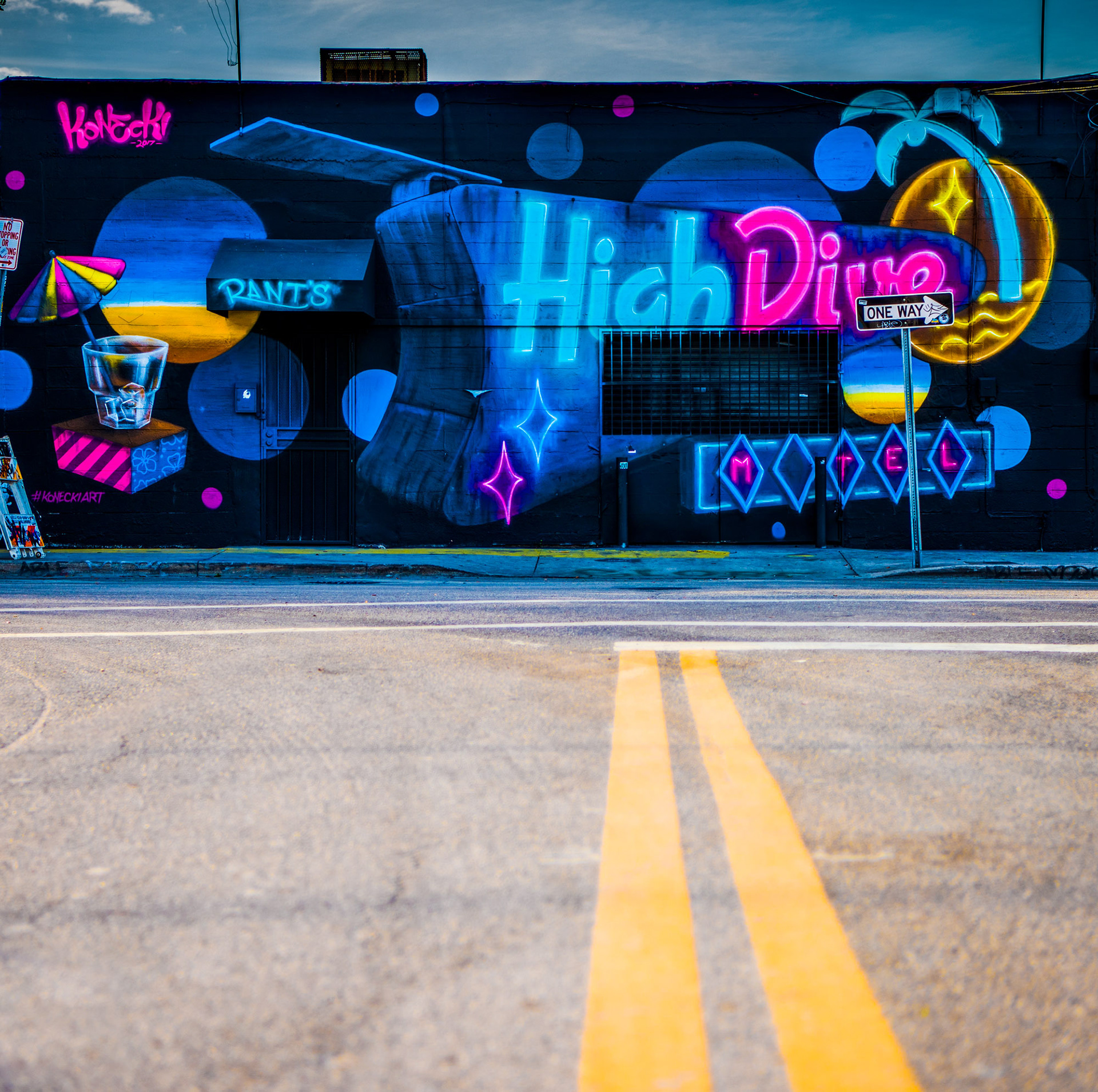Street art mural with vibrant neon colors depicting "High Dive Motel" text, tropical motifs, and geometric shapes on a dark background. Energetic tone.