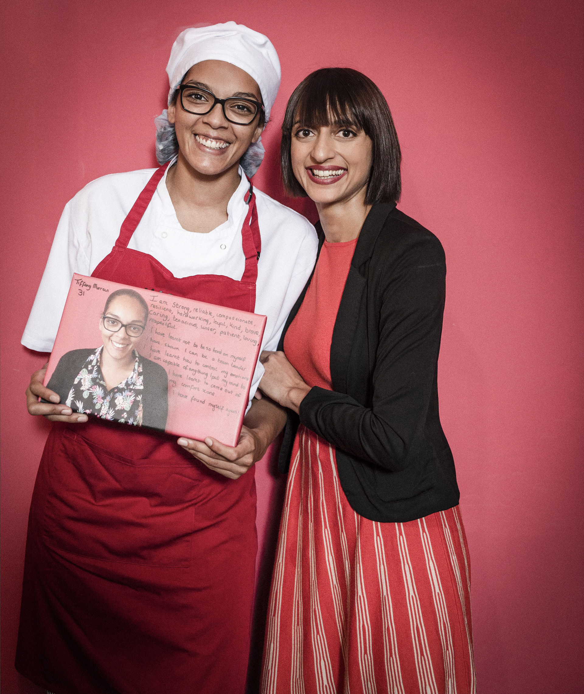 Two smiling women stand against a pink backdrop. One wears a chef's outfit, holding a poster with writing and a photo. The other wears a red dress and black blazer.