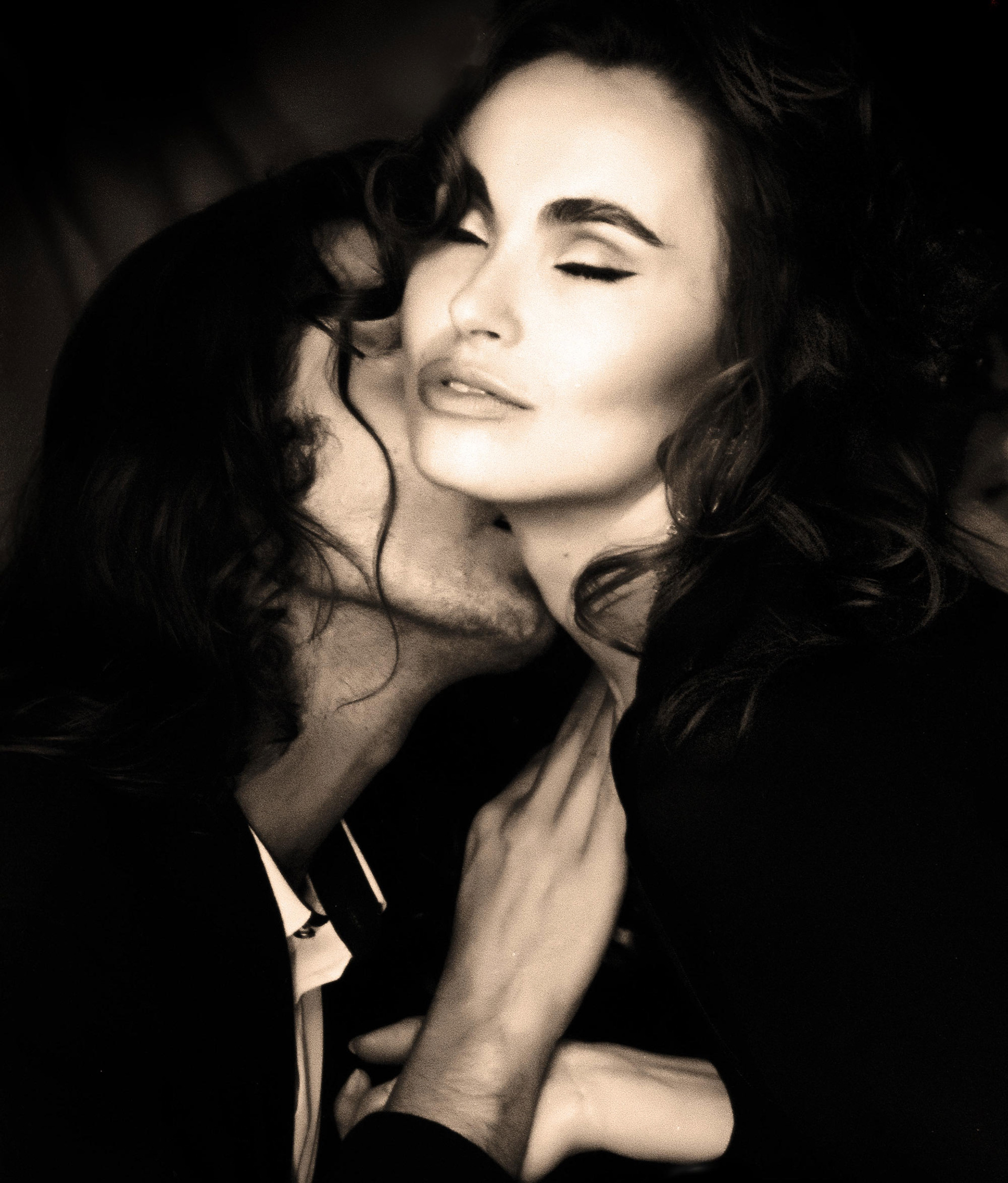 A sepia-toned image of a couple embracing intimately. The man kisses the woman's neck as she closes her eyes, conveying a sense of romance and tenderness.