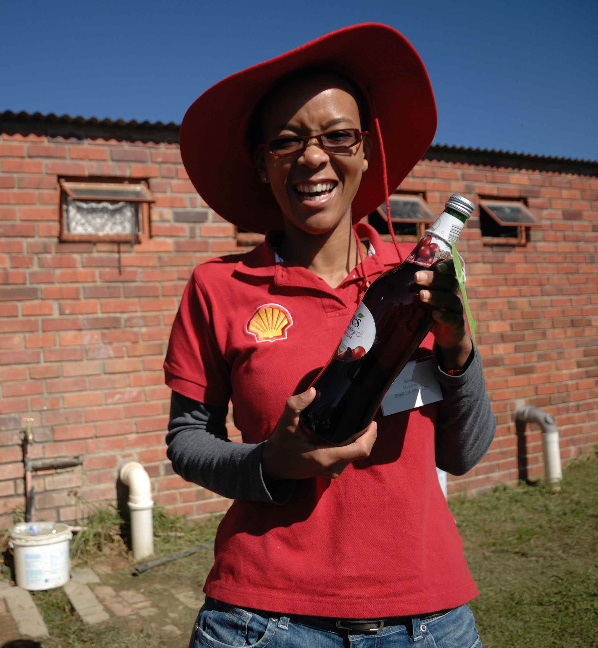 Smiling person in a red hat and shirt with a Shell logo holds a bottle of cherry juice against a brick wall backdrop under a clear blue sky.