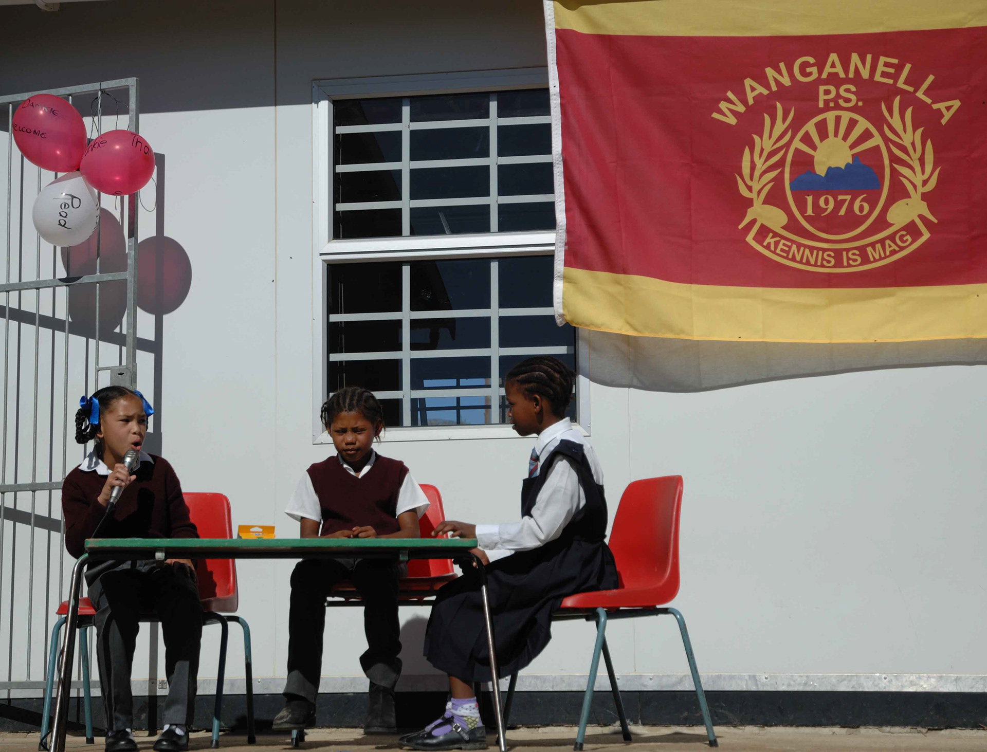 Three children in school uniforms sit on red chairs behind a table. One speaks into a microphone. Balloons and a school banner are visible behind them.