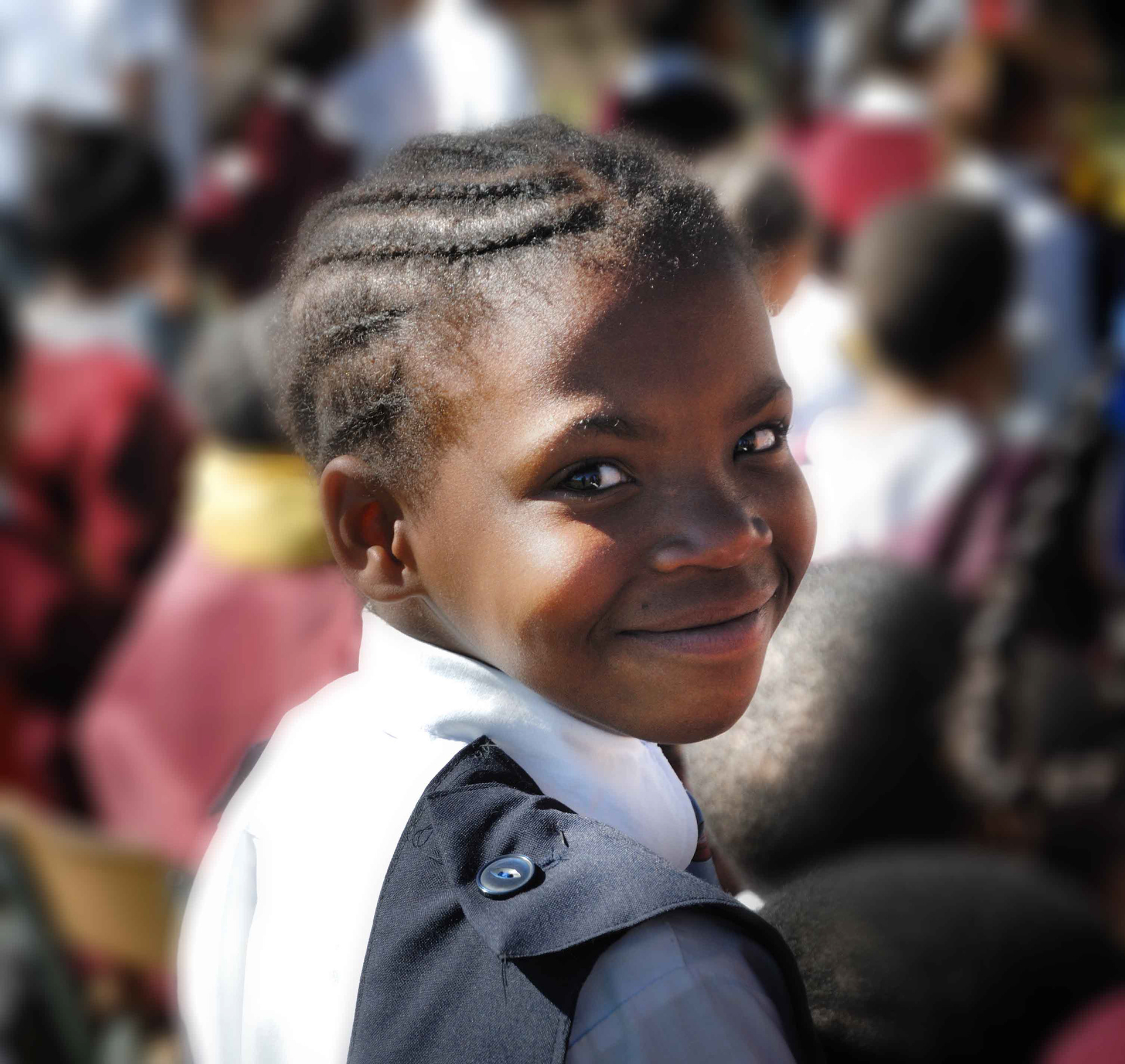 Young child smiling warmly at the camera, wearing a white shirt and dark overalls. Sunlight highlights their joyful expression, creating a positive mood.