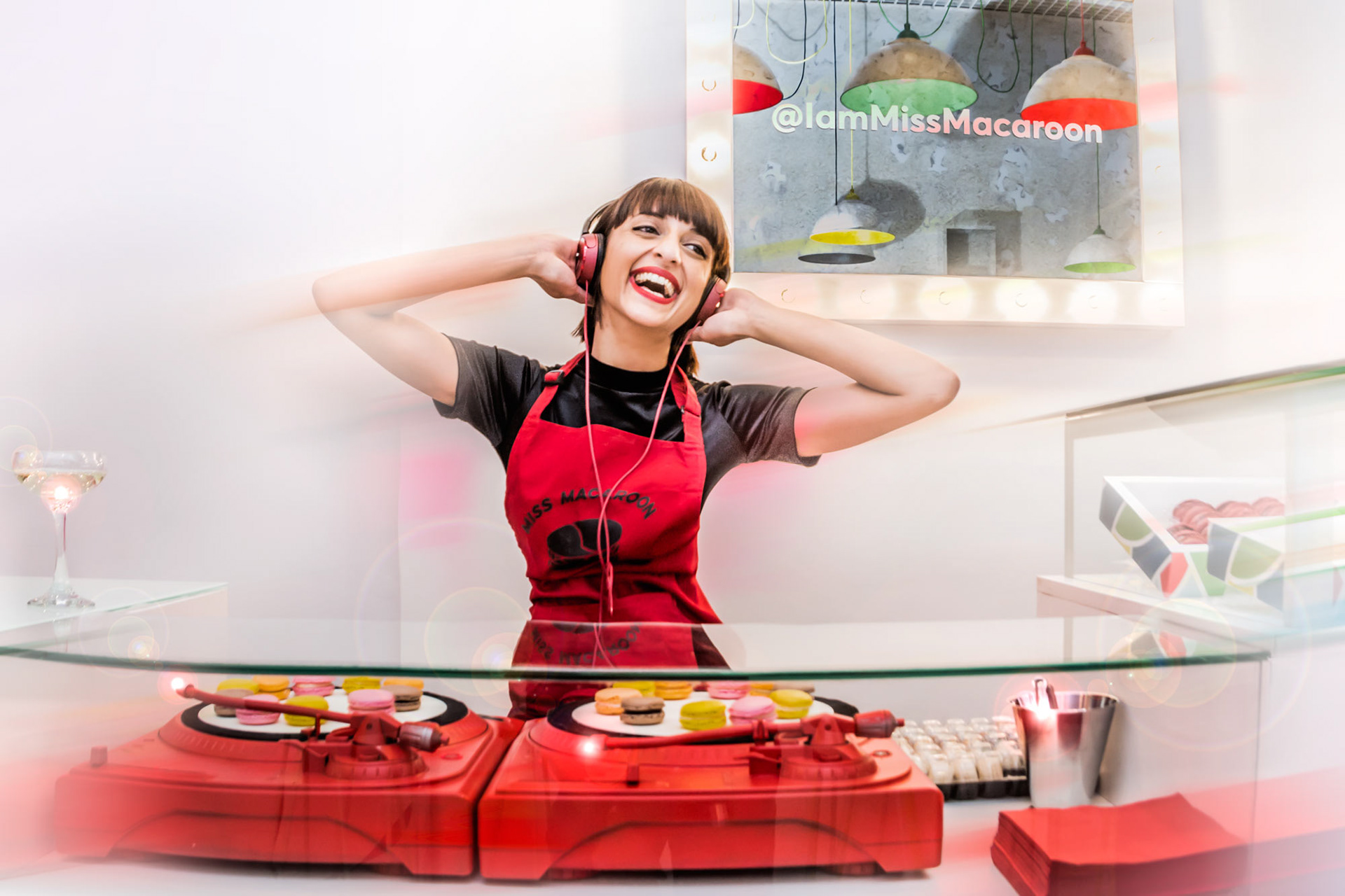 Smiling woman in a red apron wearing headphones, standing behind a DJ booth styled as a display case with colorful macarons. Atmosphere is joyful and vibrant.