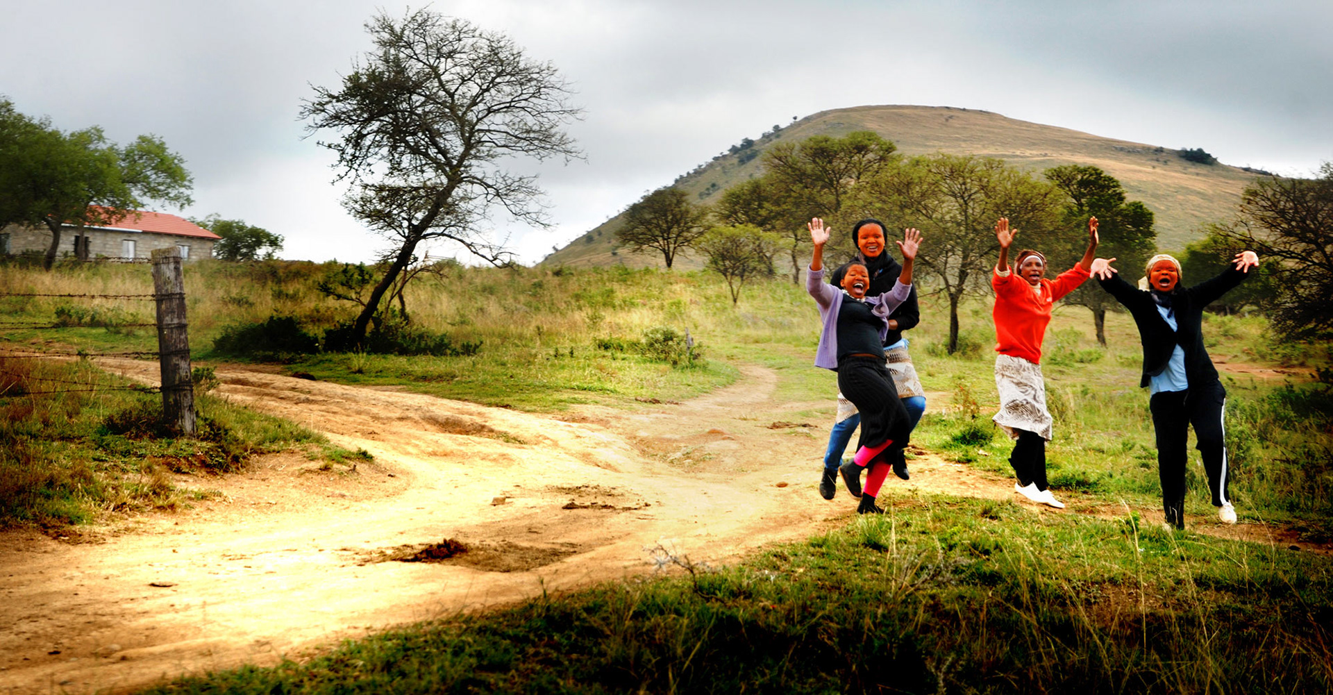 A vibrant scene of four people joyfully leaping on a dirt path in a rural landscape. Green hills and scattered trees create a lively, carefree atmosphere.