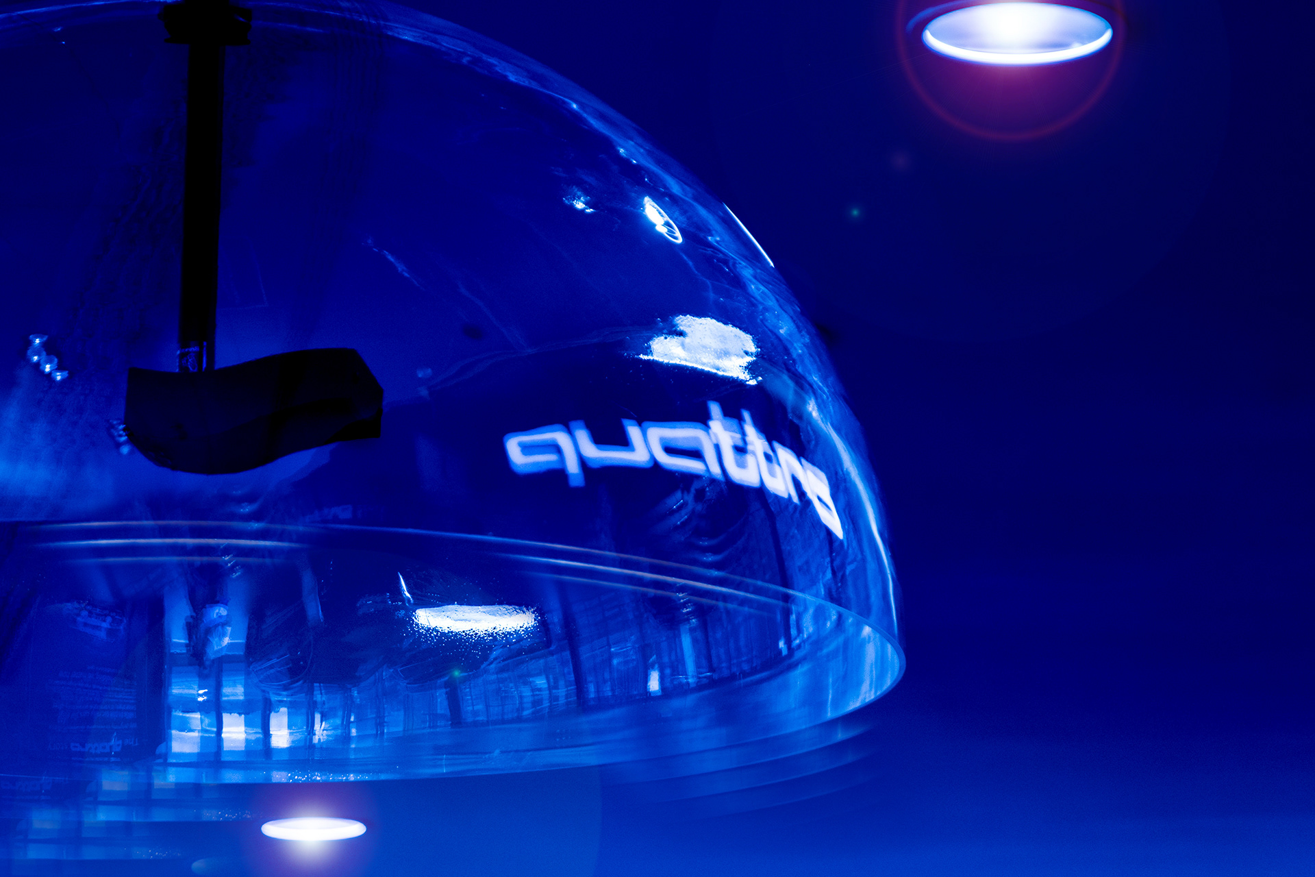 Close-up of a blue-toned, transparent bell-shaped sound dome. The word "quattro" glows on its surface. The background is blurred with bright lights.