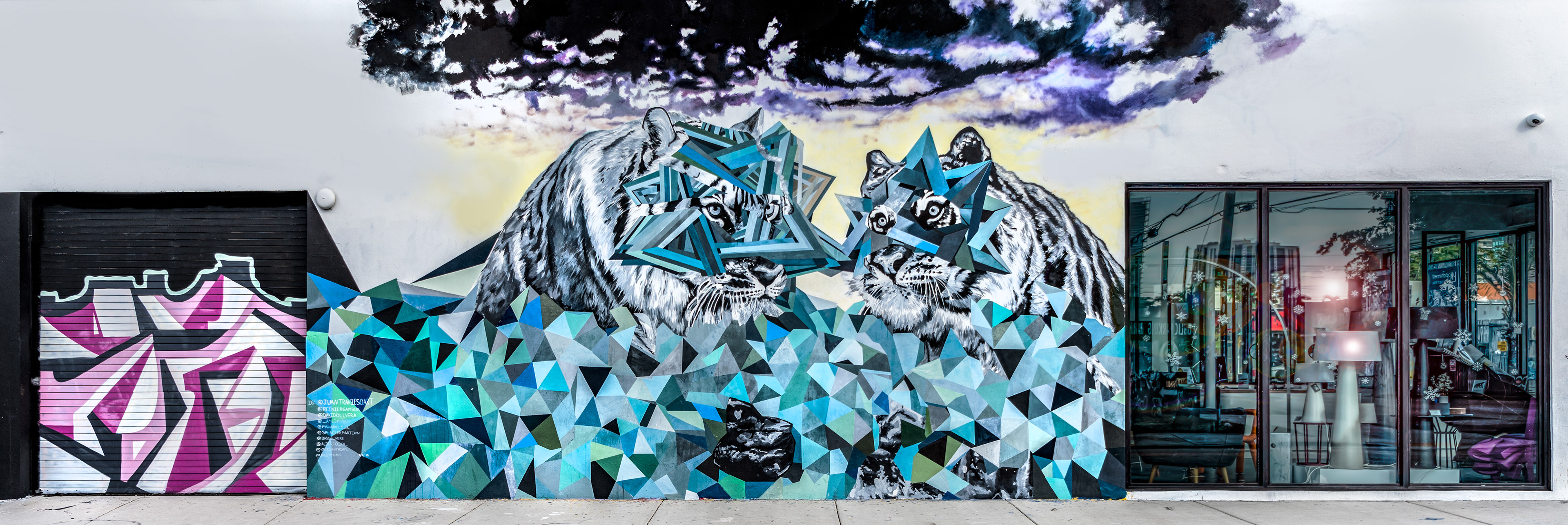 Street mural of two abstract tigers with geometric patterns and vibrant blues. Dark clouds above, modern shopfront on the right, urban atmosphere.