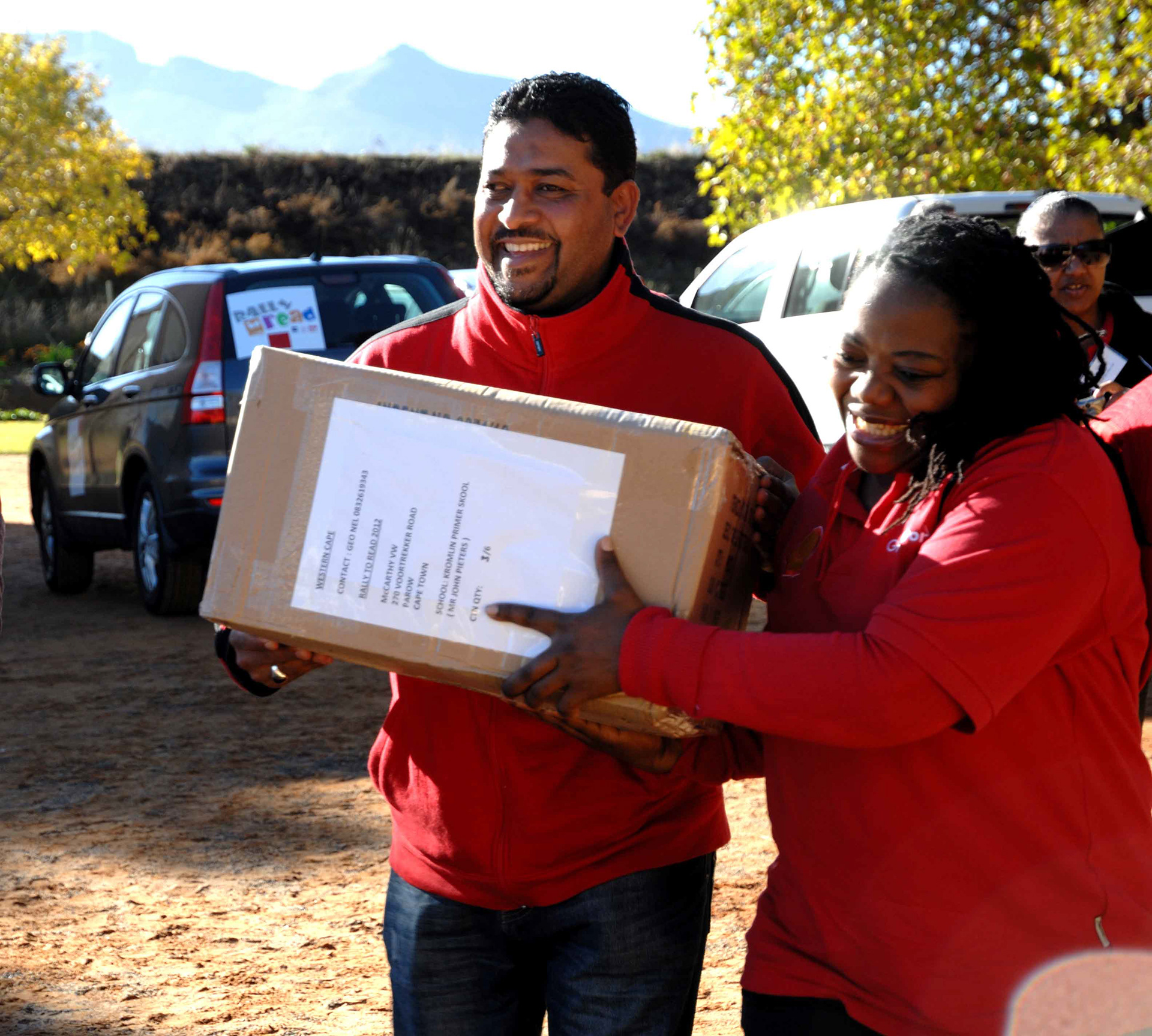 A smiling man and woman in red shirts carry a box together outdoors. Cars and trees are in the background under a clear blue sky, conveying teamwork.