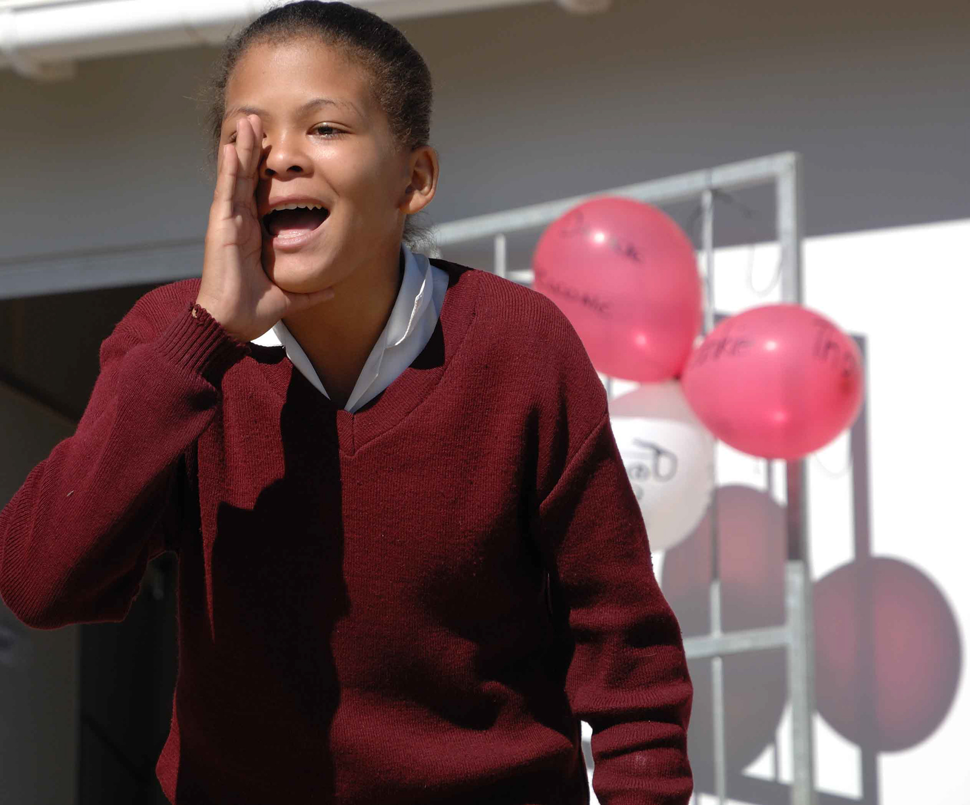 A person in a burgundy sweater energetically calls out with a hand by their mouth. Red and white balloons in the background convey a festive mood.