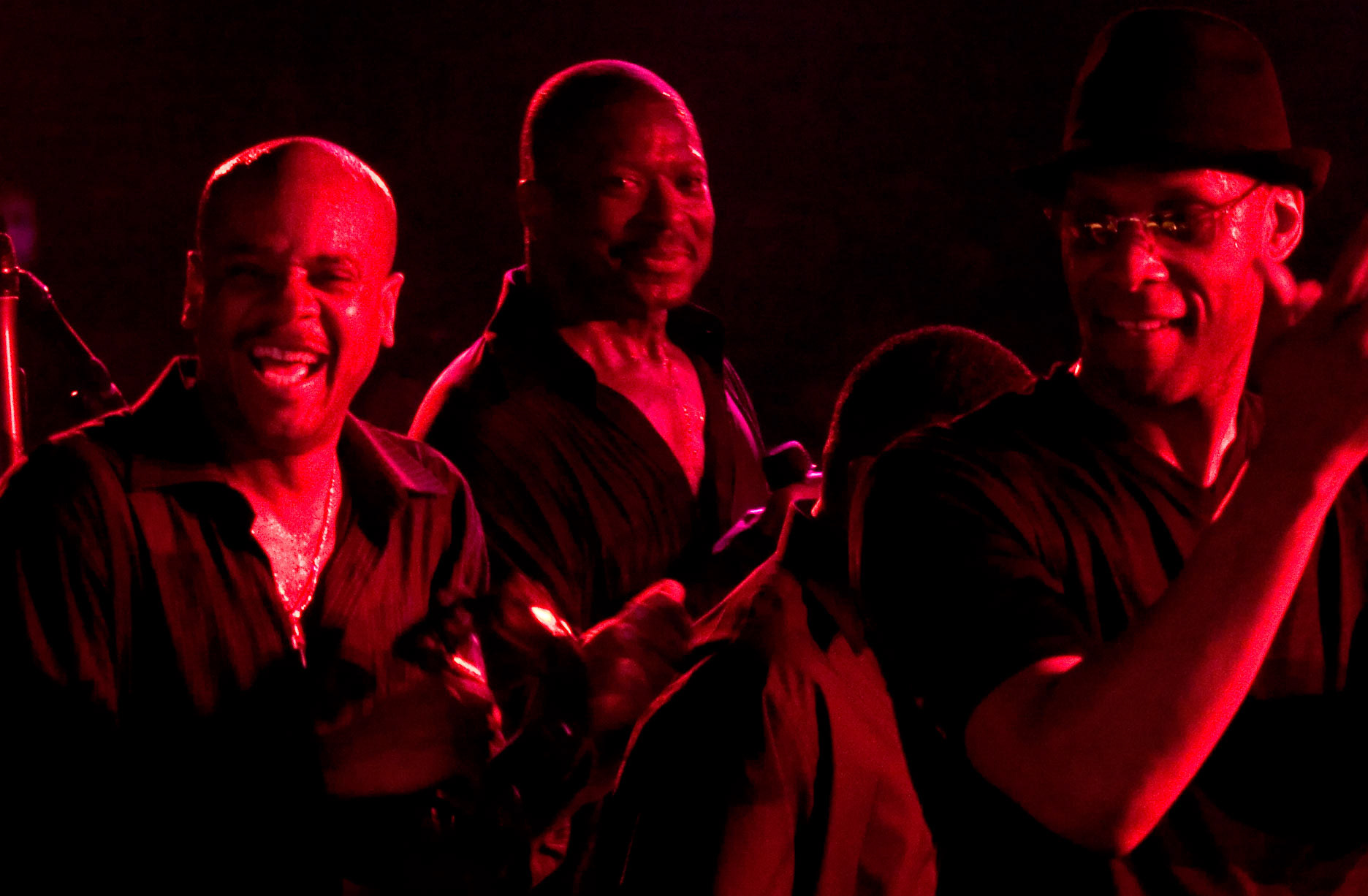 Three men in dim red lighting enjoy a lively moment, one laughing openly, the others smiling warmly. The atmosphere feels energetic and joyful.