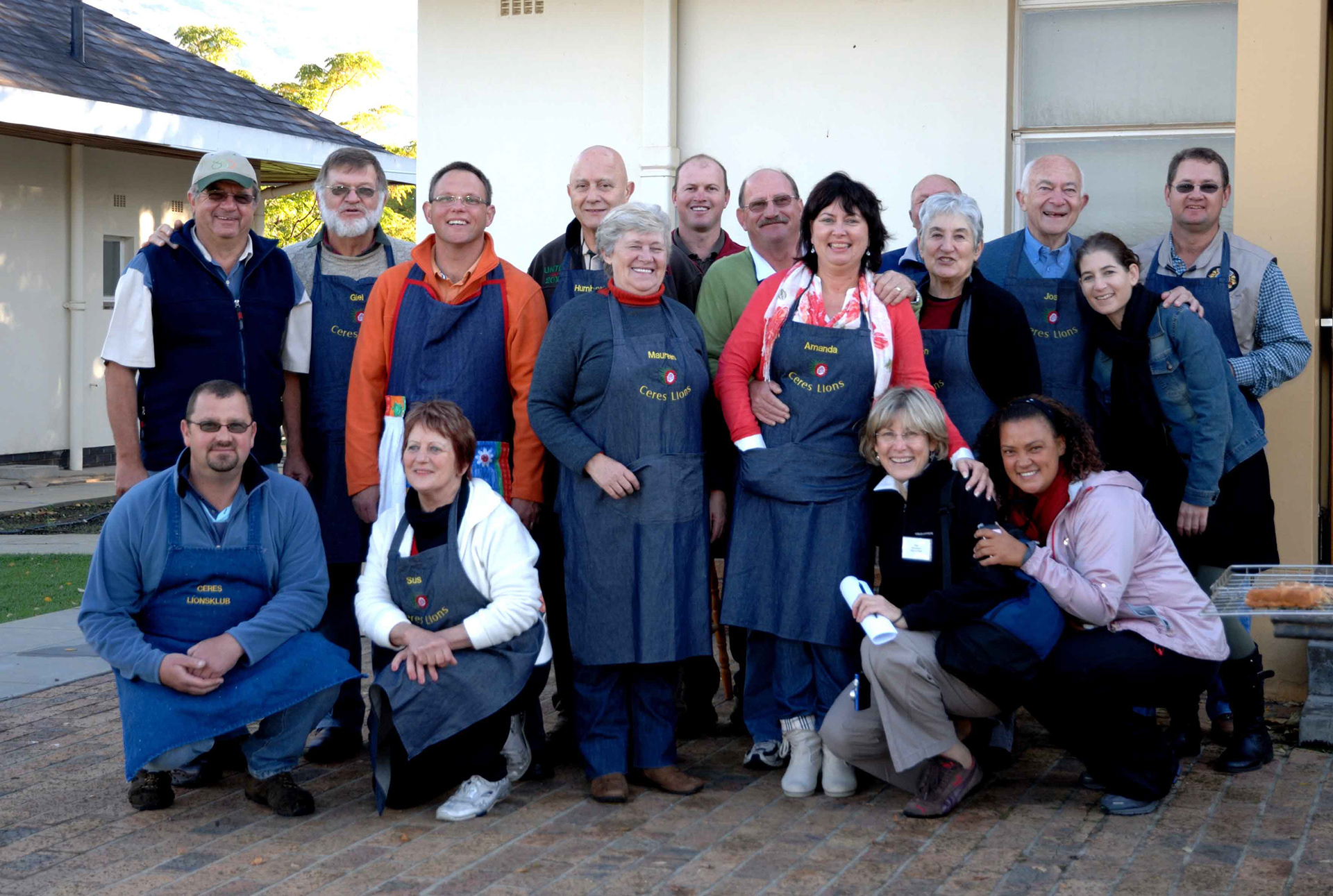 A group of 17 smiling people poses outdoors, most wearing blue aprons with "Ceres Lions" logos. The tone is friendly and community-focused.