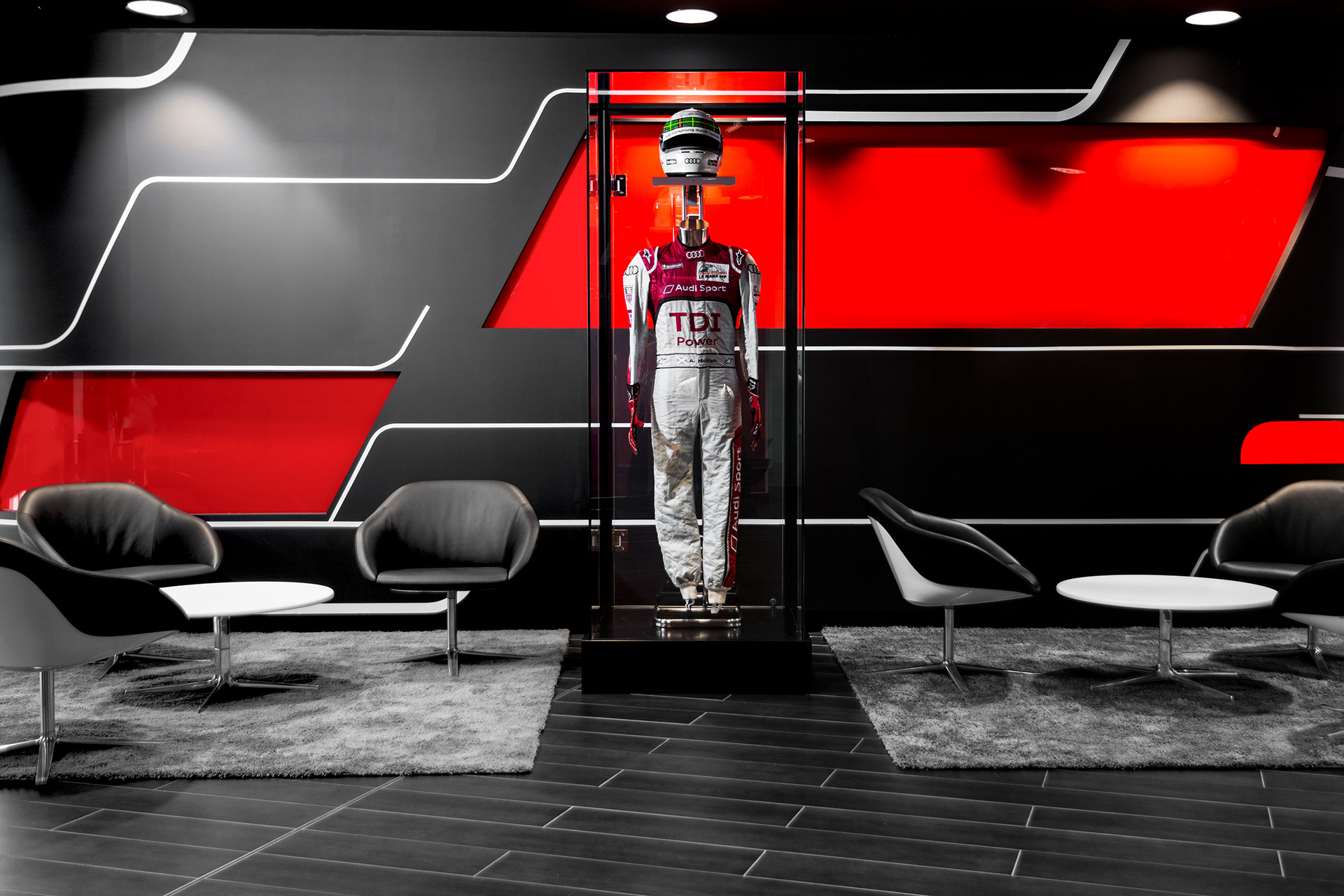 An Audi racing suit and helmet are displayed in a glass case against a sleek black and red geometric wall. Nearby, modern black chairs and white tables create a sophisticated lounge setting.
