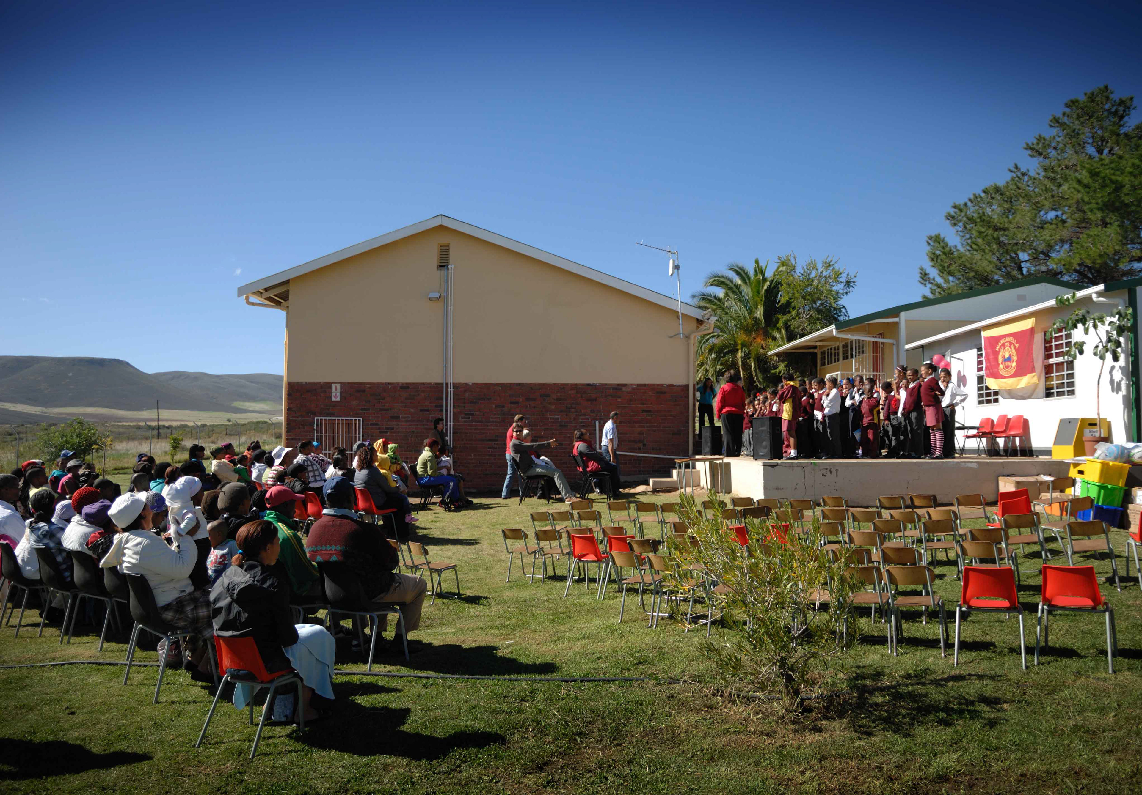 Outdoor school assembly with students in red uniforms standing on a stage, facing a seated audience under a clear blue sky. A banner is displayed.