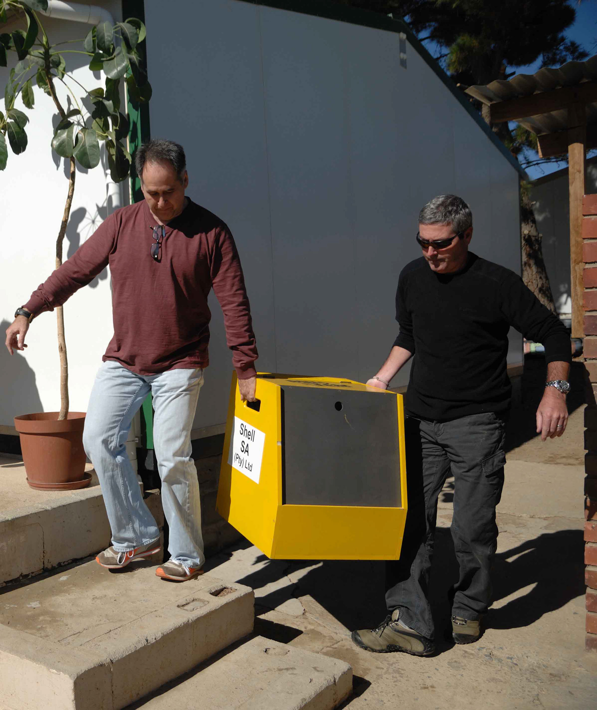 Two men carry a yellow box labeled "Shell SA (Pty) Ltd" outside on a sunny day. One wears a maroon shirt, the other black, suggesting teamwork.