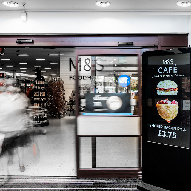 Store entrance of M&S Foodhall. A digital sign advertises a café's smoked bacon roll for £3.75. Blurred figures walk inside, creating a busy atmosphere.